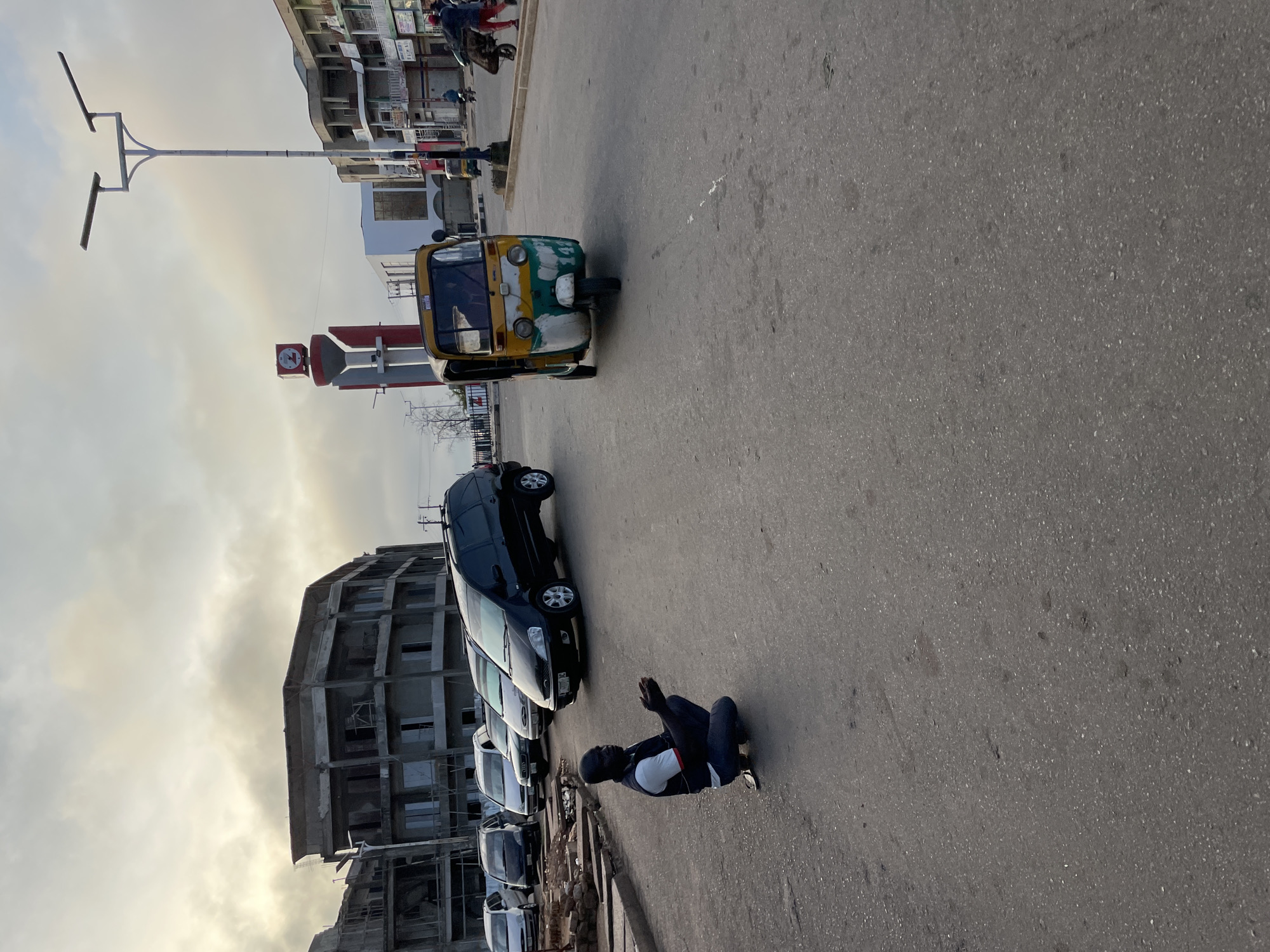 A man kneels on the asphalt road with his hands clasped in supplication. He wears a dark vest over a white t-shirt and dark pants. Behind him, several cars are parked, and a black minivan is in motion. To the right, a yellow and green auto-rickshaw navigates the street. In the background, buildings under construction and a clock tower signify the urban environment of Jos, Nigeria. The sky is overcast, suggesting a cloudy day.