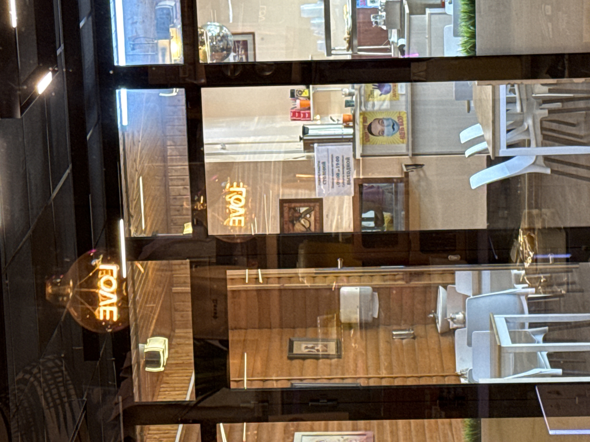 The image captures a reflection of a cafe interior seen through a glass storefront. A prominent neon sign spelling "LOVE" in Cyrillic, with the letters backwards, is illuminated within a large glass bulb hanging from the ceiling. The cafe features wooden paneling on the walls and a seating area with white tables and chairs. In the background, a poster of a person wearing a face mask with the text "НАДЕНЬ МАСКУ" (Wear a mask) is visible. Another poster shows a hand with the text "ЗАПРЕЩЕНО" (Forbidden). The overall scene suggests a casual dining establishment, likely a cafe or restaurant, with a subtle nod to public health guidelines. The presence of the "LOVE" sign could indicate a romantic theme or simply a decorative element of the establishment. The reflection in the glass also shows a disco ball and part of a tiled ceiling.