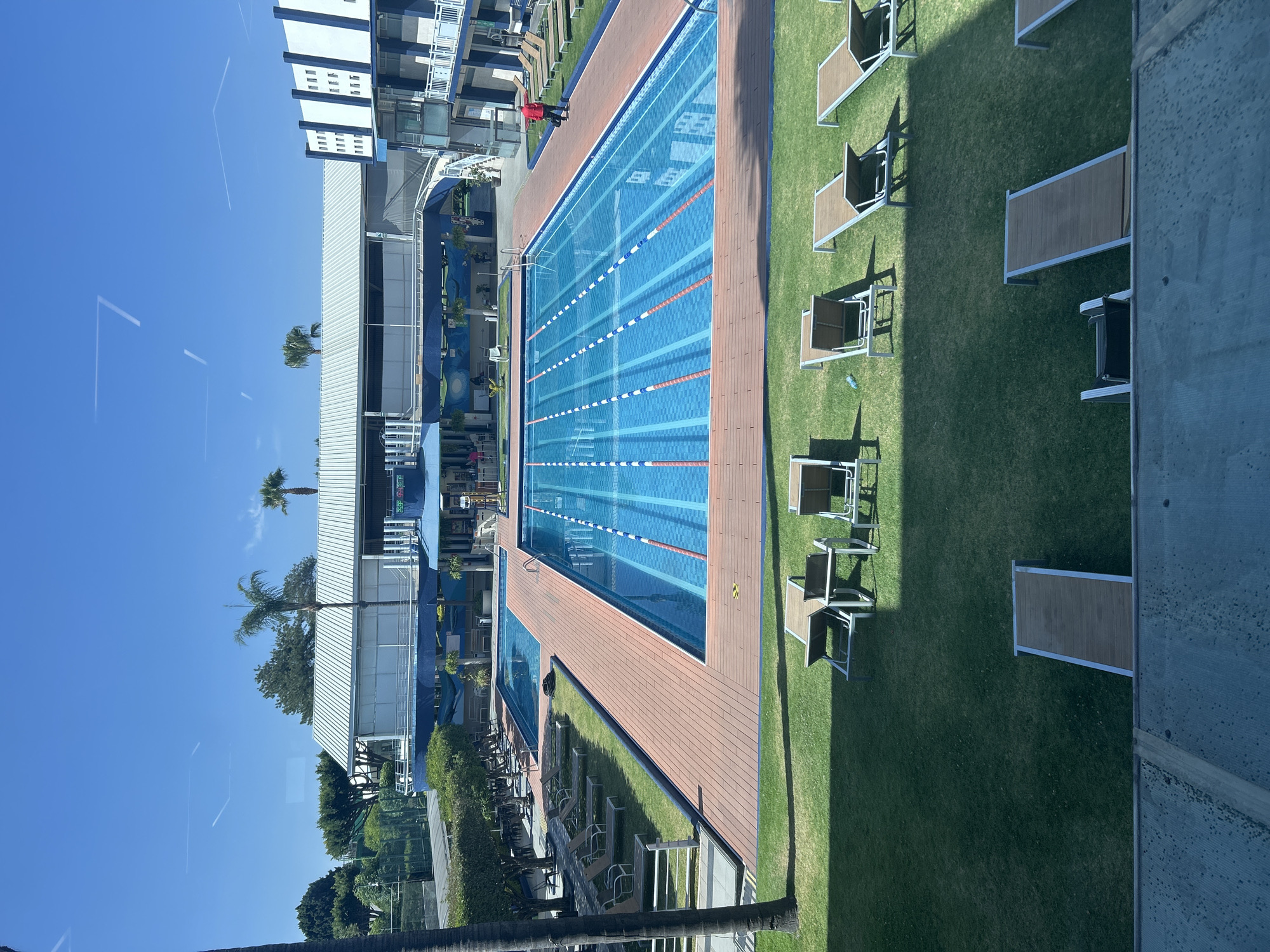 The image depicts a swimming pool complex on a sunny day. The complex features a large rectangular pool with multiple lanes, delineated by lane markers. The pool is surrounded by a wooden deck and a grassy area with lounge chairs. A multi-story building is located adjacent to the pool area. The blue sky is clear, with some palm trees visible in the background. A person dressed in red is present, walking by the pool. The pool is located in Puebla, Mexico.