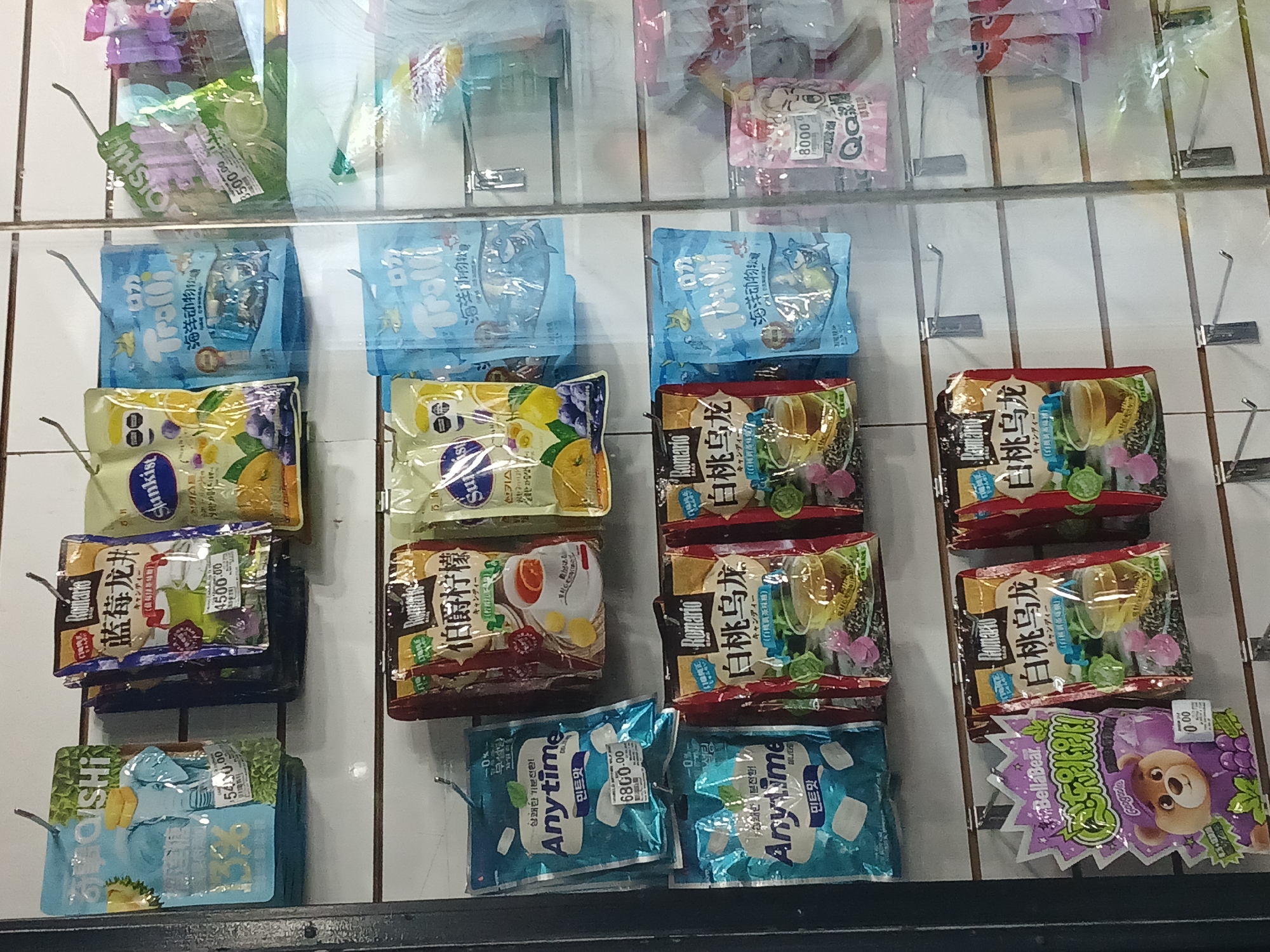 This image captures a retail display cabinet filled with various packaged candies and snacks, likely from an Asian market in Buenos Aires, Argentina. The products are neatly hung on metal hooks across multiple shelves behind a transparent barrier, suggesting a vending machine or a store display.
The subjects are numerous colorful packages of confectionery. Brands visible include "OISHI", "Trolli" (featuring marine animal-shaped candies, labeled "海洋动物"), "Sunkist" (depicting fruits like lemon, orange, and grape), "Romario" (with flavors such as "伯爵柠檬" or Earl Grey Lemon, and "白桃乌龙" or White Peach Oolong tea candies), "Anytime" (mint-flavored lozenges, labeled "민트맛" and "상쾌한 기분전환" for a refreshing change of mood), "BellaBear", and "Q&Q" (one package showing "草莓味" or strawberry flavor).
Notable details include the multilingual packaging, primarily featuring Chinese, Korean, and Japanese text, alongside English brand names. Several packages display numerical values, likely prices, such as "500.00", "4500.00", "5400.00", "6800.00", and "8000", with some showing "0.00". The presence of these high numerical values for individual items is consistent with prices in Argentine Pesos. The overall impression is that of a diverse selection of international snacks available for purchase. The setting is indoors, with no indication of the time of day or weather.