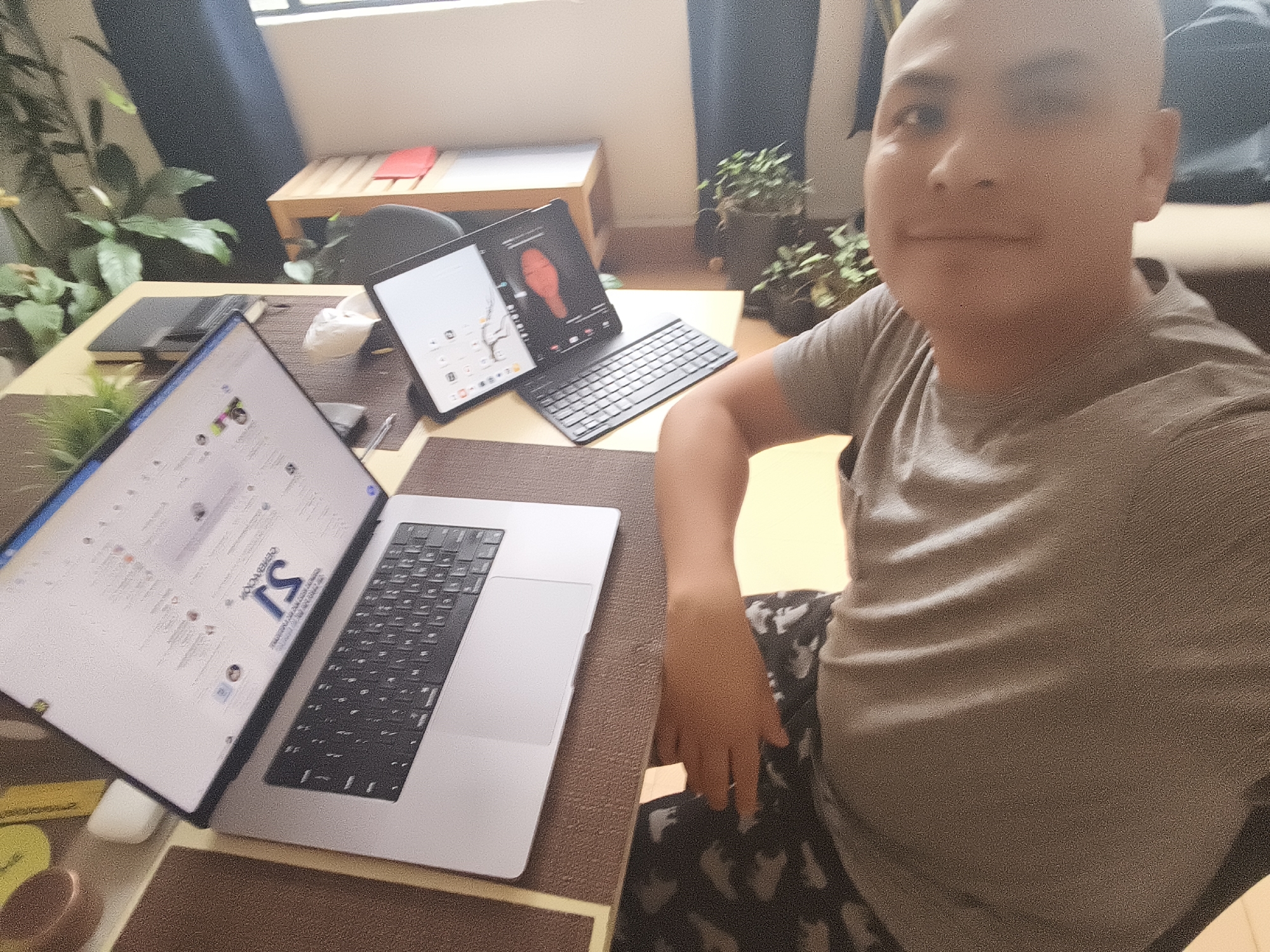 A bald person in a gray shirt is seated at a desk. On the desk is a laptop displaying a document with the letters "TS" in bold, and a tablet with a connected keyboard. Other items on the desk include a notepad, a pen, a mouse, and small containers. Plants are visible in the background. The location is inferred to be Mexico City, Mexico.