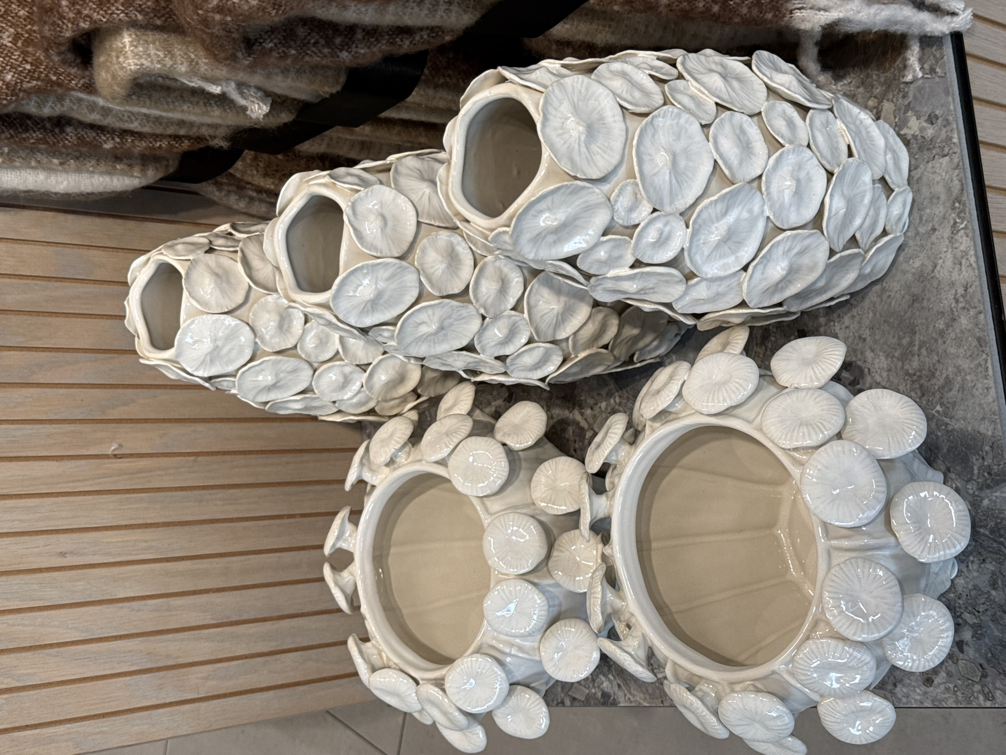 The image shows four white, ceramic vases with a textured surface of circular disks. Two vases are elongated with multiple openings, while the other two are bowl-shaped. They are placed on a gray surface, possibly a table or shelf. A portion of a folded textile with brown and black patterns is visible in the upper left corner. The background includes a wall with horizontal wooden planks. The scene is well-lit and the objects appear to be indoors. The location in Cape Town, South Africa, is not evident from the visual information provided.