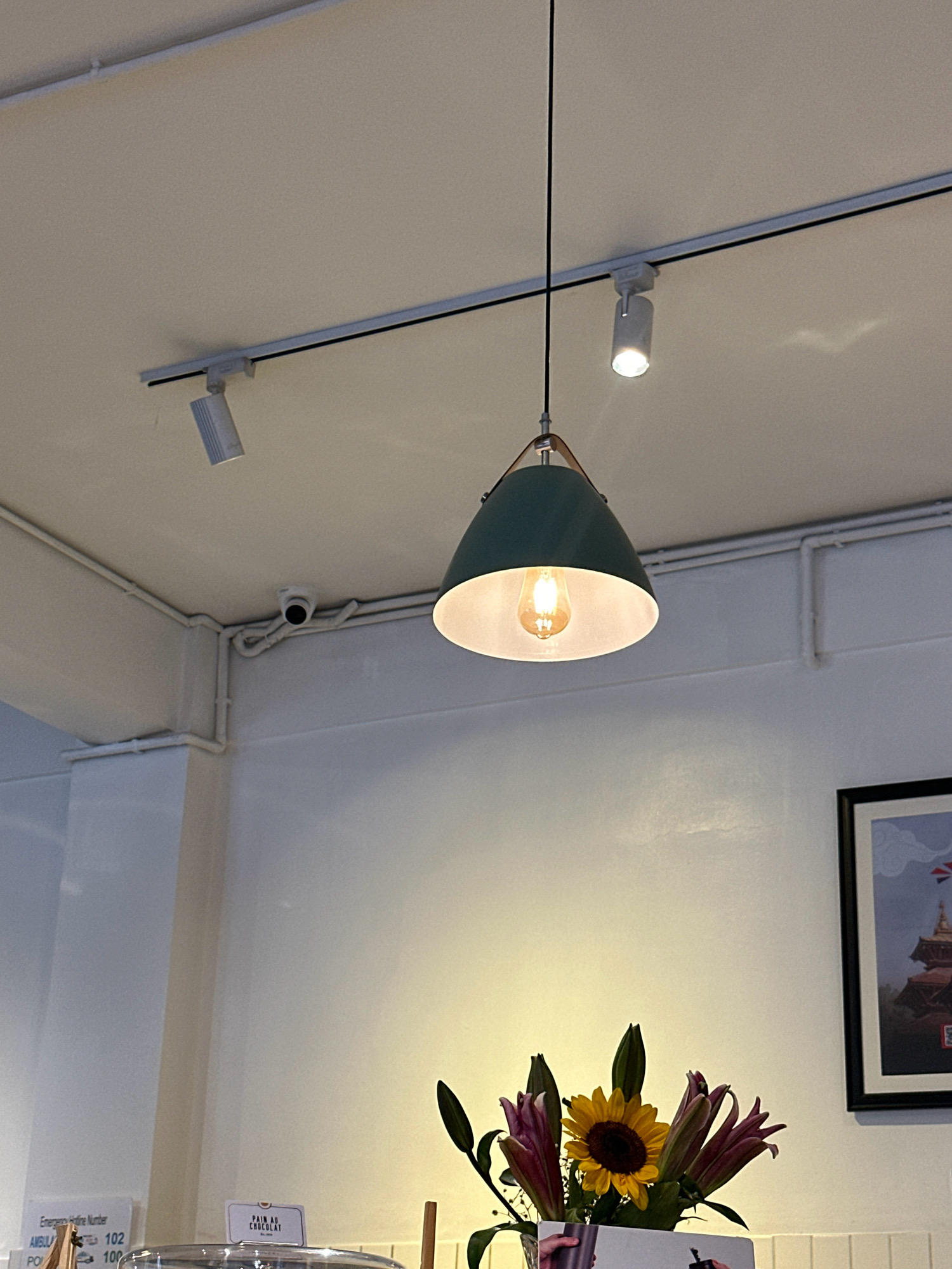 The image captures an indoor scene, likely a cafe or shop, with a white ceiling and walls. A track lighting system is installed on the ceiling, with several adjustable spotlights. A pendant light with a teal lampshade hangs from the track, featuring an Edison-style light bulb. A small security camera is visible on the ceiling near the corner. In the foreground, a bouquet of flowers, including a sunflower and lilies, is arranged in a vase. To the left of the flowers, there's a display with small signs, one indicating "PAIN AU CHOCOLAT." A framed picture is mounted on the wall to the right, depicting a pagoda-like structure. The overall ambiance suggests a well-lit, modern interior. The scene does not contain any identifiable individuals or specific activities. The location of Lalitpur, Nepal, is not explicitly depicted or suggested by any visual cues in the provided image.