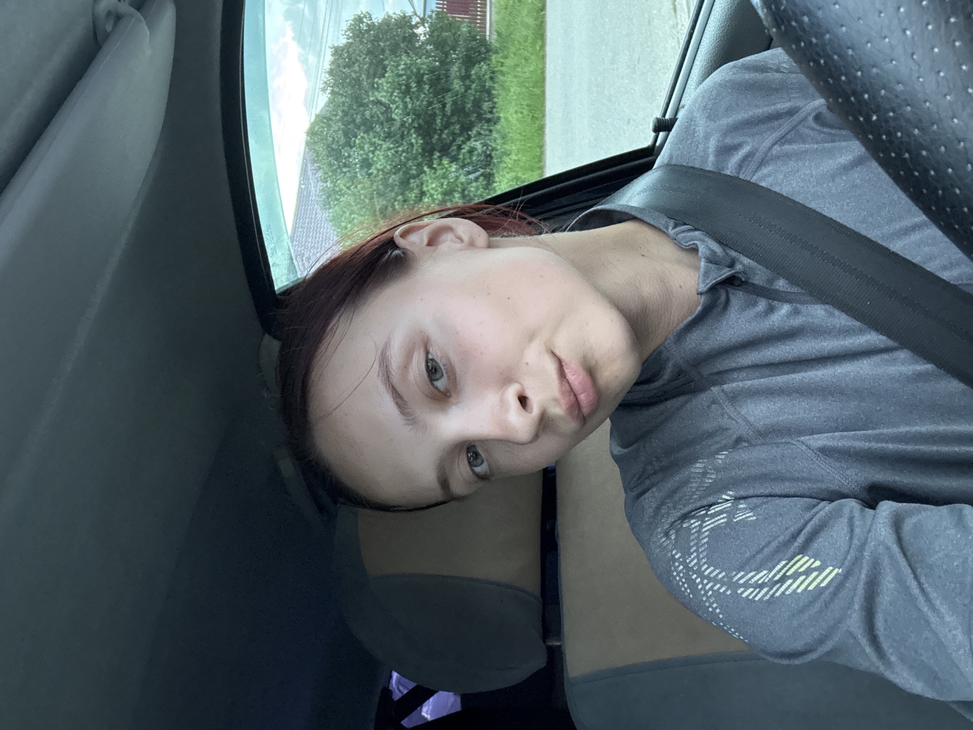A young adult female is depicted in a selfie-style photograph taken inside a vehicle, located in the city of Прикамский, Russia. She is seated in the driver's position, with a black steering wheel partially visible in the lower right corner, and is secured by a black three-point seatbelt across her left shoulder and chest. Her hair is dark reddish-brown and pulled back, and her eyes are light green-blue. Her facial skin is fair with light freckles, and her lips are slightly puckered. She is wearing a grey long-sleeved athletic shirt with a quarter-zip collar, featuring a white and light green abstract geometric pattern on the left shoulder. The vehicle's interior headliner and trim are grey, and a tan and grey car seat headrest is visible behind her. Through the car's right-side window, a daytime outdoor scene is visible, including a large green deciduous tree, a red-painted wooden fence, green grass, and a light-colored sky with some cloud cover. The car window exhibits a slight blue-green tint.