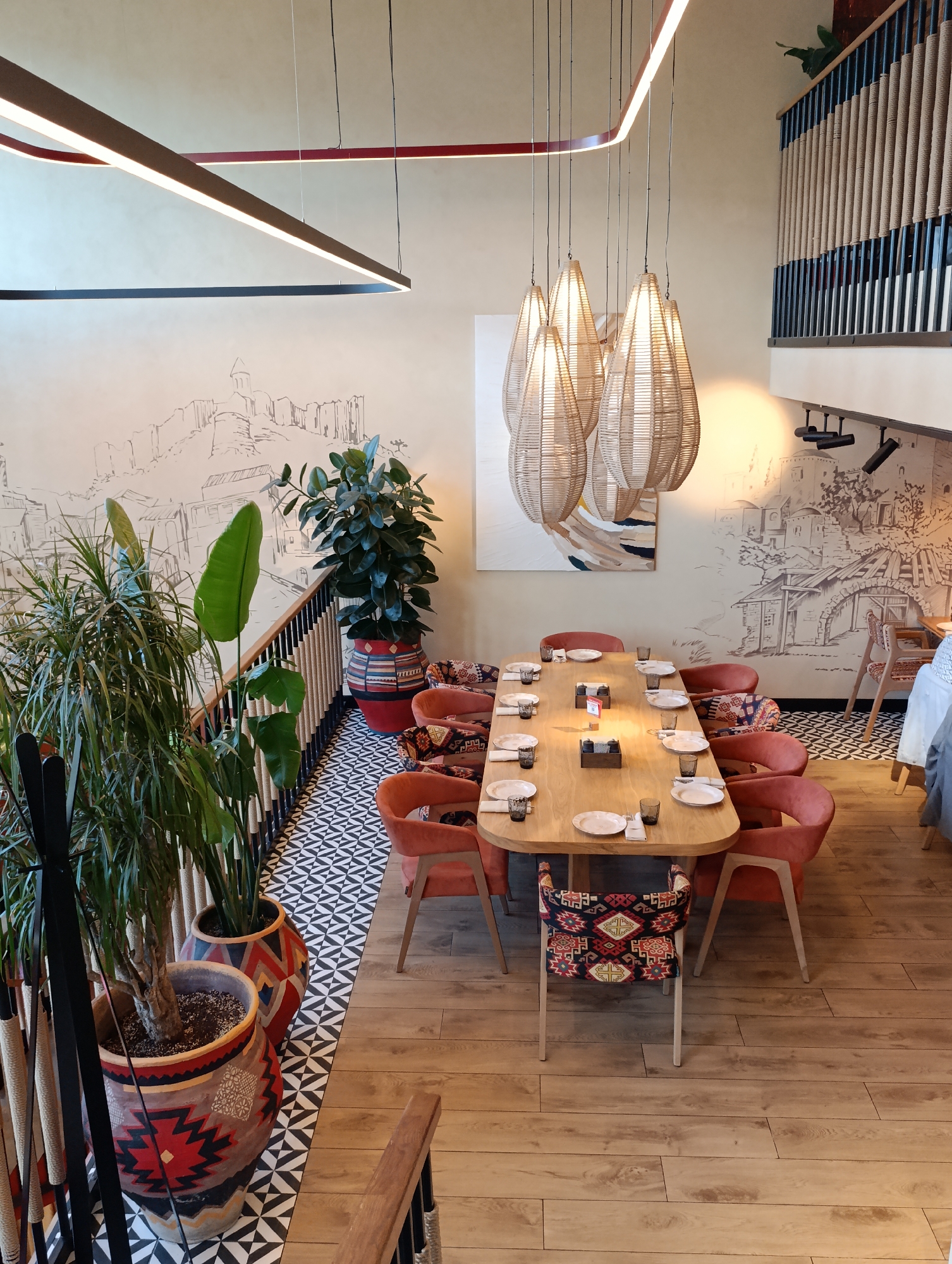 The image displays the interior of the Tsakhkadzor restaurant, located at Bolshoy Gnezdnikovskiy Pereulok, 10, Moscow, Russia. The perspective is from an elevated vantage point, looking down into a main dining area and across to an upper-level balcony.
The ceiling features multiple suspended linear LED light fixtures; a large, dark rectangular frame is prominent, while a reddish-brown curved segment is visible higher up. A cluster of five tear-drop shaped pendant lamps, constructed from woven material, hangs above the main dining table. The walls are a light beige, adorned with extensive monochromatic murals depicting a detailed, stylized landscape of mountains, old town buildings, and a bridge. Track lighting on the far wall illuminates a section of the mural.
The primary dining area is floored with light-colored wooden planks. Adjacent to a railing, a section of black and white geometric patterned tiles is present. The railing itself is constructed from dark metal vertical bars interwoven with thick, light-colored rope or cord. A similar railing is visible on the upper-level balcony in the background.
A large, oval-shaped wooden dining table, light in tone, is centrally positioned and set for multiple guests, with white plates, dark glasses, and cutlery at each of the ten visible settings. Small wooden boxes are arranged along the table's center. The chairs comprise two styles: solid reddish-orange/terracotta upholstered chairs with light wooden frames, and chairs upholstered with vibrant, geometric-patterned fabric in shades of red, black, beige, and green.
Several large, green potted plants are strategically placed throughout the space. These include tall plants with spiky leaves and broad-leafed plants, contained within large ceramic pots decorated with colorful, geometric patterns in red, black, white, and orange. An abstract artwork, featuring light blue, brown, and cream tones, hangs on the far wall above a partially visible dining setup with a white tablecloth and a light blue fabric draped over a chair. No individuals are present in the scene.