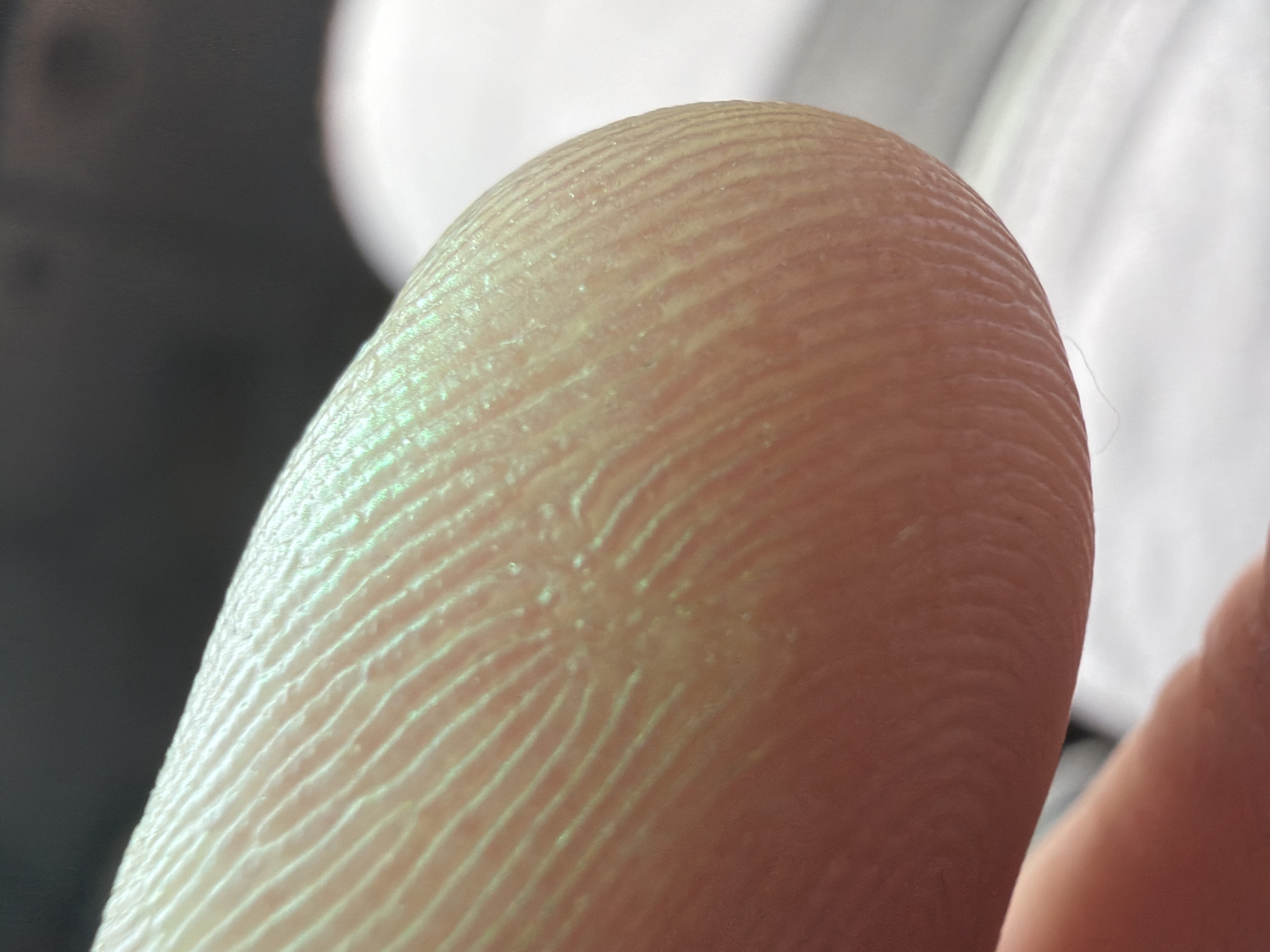 The image is an extreme close-up of a human fingertip. The subject is a person's finger, with visible fingerprint ridges. The skin appears pale with a slightly yellowish or greenish tint due to the lighting. The background is out of focus, with a dark, shadowed area on the left and a blurred bright area on the right, suggesting artificial lighting or a bright light source out of frame. There is a hint of another finger or hand in the lower right corner, also out of focus. The activity appears to be a static shot, focusing on the texture of the skin and the detail of the fingerprint. There are no visible location cues, time of day indicators, weather conditions, or emotions conveyed. No text is visible in the image.