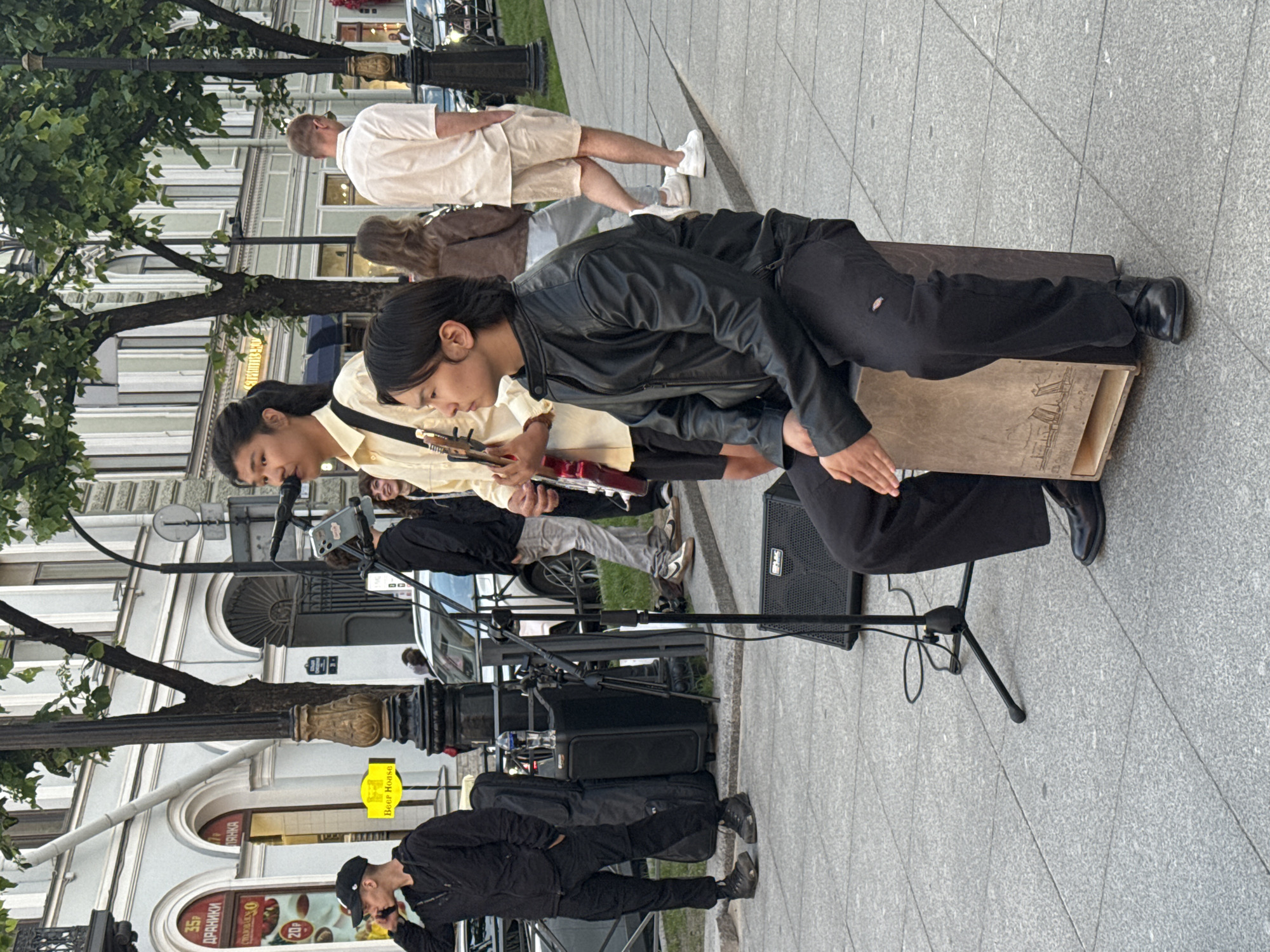 The image depicts a street scene in Saint Petersburg, Russia, likely on a daytime with overcast skies. In the foreground, two young men are performing music on a sidewalk. One man, seated on a cajon drum and wearing a black leather jacket and black pants, is looking to the left. The other man, standing and wearing a light yellow shirt, is playing a red electric guitar and singing into a microphone. He has a guitar strap over his shoulder.
Behind the performers, a black speaker sits on a tripod, and a black guitar case is leaning against a lamp post. Other people are visible in the background, including a man in a black hoodie and baseball cap talking on his phone, and several individuals walking along the street. Buildings with classical architecture, characteristic of Saint Petersburg, line the background. The sidewalk is paved with gray granite tiles. The overall atmosphere suggests a casual outdoor performance in an urban setting.