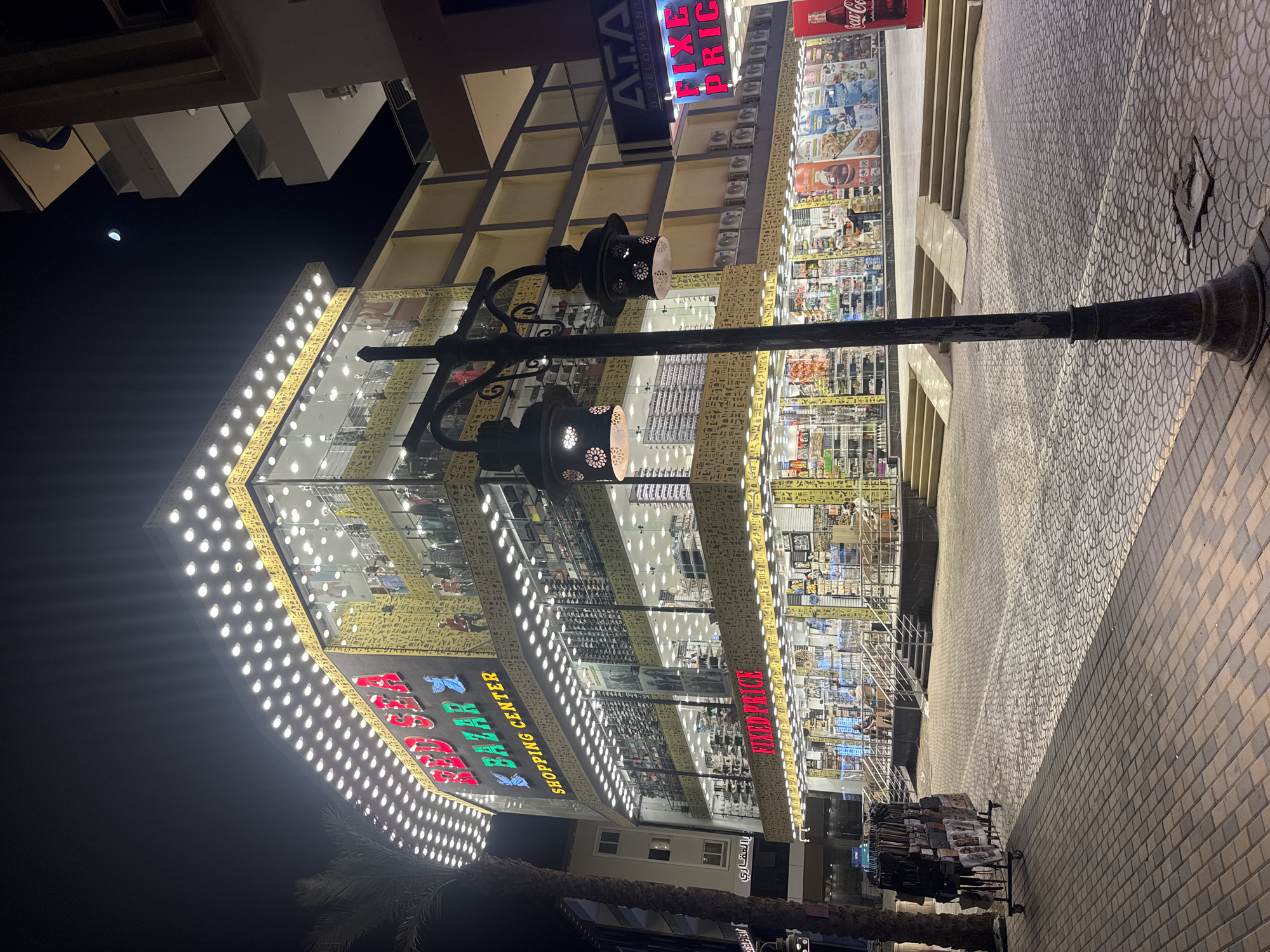 A nighttime shot captures the "Bazar Red Sea Shopping Center" in an unknown city. The building's facade is adorned with rows of lights and Egyptian hieroglyphs, illuminating its multi-story structure filled with various goods. A black lamppost with decorative lamps stands in the foreground. The street pavement, with its patterned brickwork, extends into the building. The moon can be seen faintly in the dark night sky.