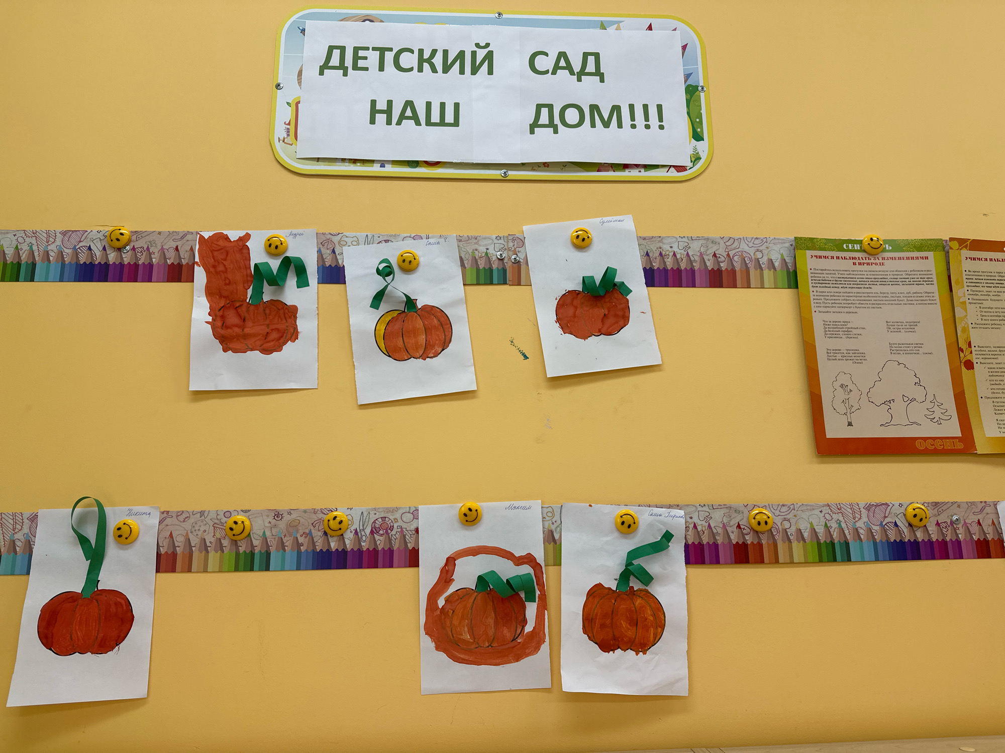 The image is a photograph taken inside a room with yellow walls. At the top, a sign reads "ДЕТСКИЙ САД НАШ ДОМ!!!" which translates to "Kindergarten Our Home!!!" The sign is framed by a colorful border with cartoon characters.
Below the sign, there are several pieces of children's artwork displayed on the wall, attached with yellow smiley face magnets. The artworks primarily depict pumpkins, painted in various shades of orange. Some of the artworks also have green paper leaves or stems attached.
On the right side of the image, there are two educational posters. One poster has the title "СЕН" which is likely the beginning of a word. Below the title, there are bullet points with text. The other poster, partially visible, also contains text and illustrations of trees, with the word "Осень" (Autumn) at the bottom.
The overall scene suggests a classroom or a common area in a kindergarten, possibly decorated for an autumn theme given the pumpkin artwork and the "Autumn" poster. The image does not contain any identifiable landmarks or details that would allow for a precise location within Vidnoye, Russia, beyond it being an indoor setting. There are no people visible in the image.