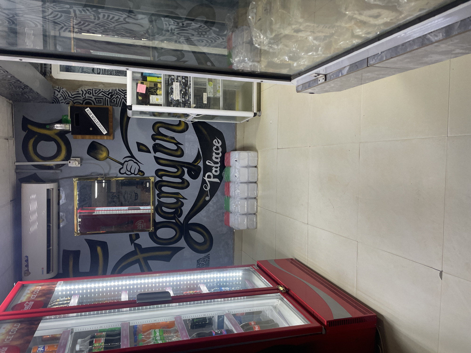 The image depicts the interior of a small convenience store, likely in Barako, Nigeria. On the left, a large, red, glass-door refrigerator is filled with various beverages, including Coca-Cola bottles. In the background, the wall features a mural with stylized lettering and decorative patterns. A gold-framed mirror hangs on the wall, reflecting a portion of the store. Above the mirror, an air conditioning unit is mounted. To the right of the mirror, a wooden suggestion box with a "SUGGESTION BOX" label is attached to the wall. In the center of the image, a white display cabinet contains shelves stocked with packaged goods. In front of the display cabinet, a row of plastic water bottles with colored lids are neatly arranged on the tiled floor. The floor itself is tiled with light-colored squares. The overall scene suggests a commercial establishment that sells drinks and possibly other convenience items.