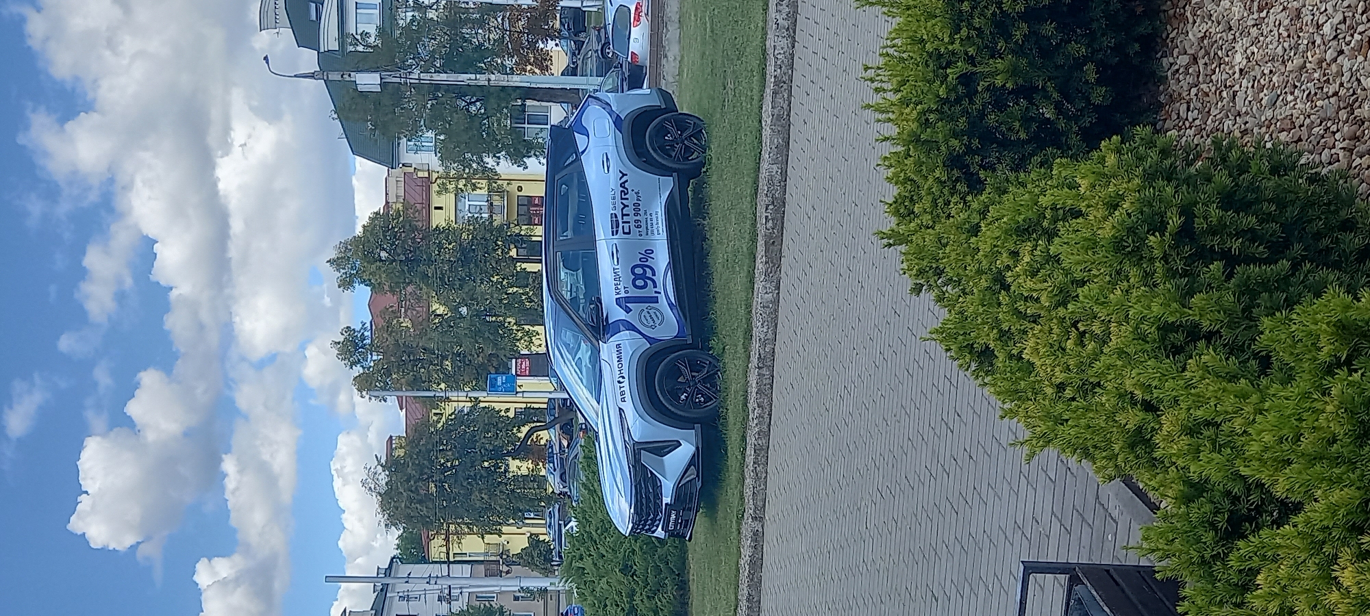 A silver SUV with blue and white graphics advertising a car loan at 1.99% interest is parked on a grassy area. The car is branded "GEELY CITYRAY" and "АВТОНОМИЯ". The text on the graphic reads "КРЕДИТ от 1,99%" and "CITYRAY от 69 900 руб.". In the background, there are trees and buildings, indicating an urban setting. To the left of the car, a paved walkway is visible, bordered by green bushes. The sky is partly cloudy. The location is identified as Brest, Belarus, based on the provided information.