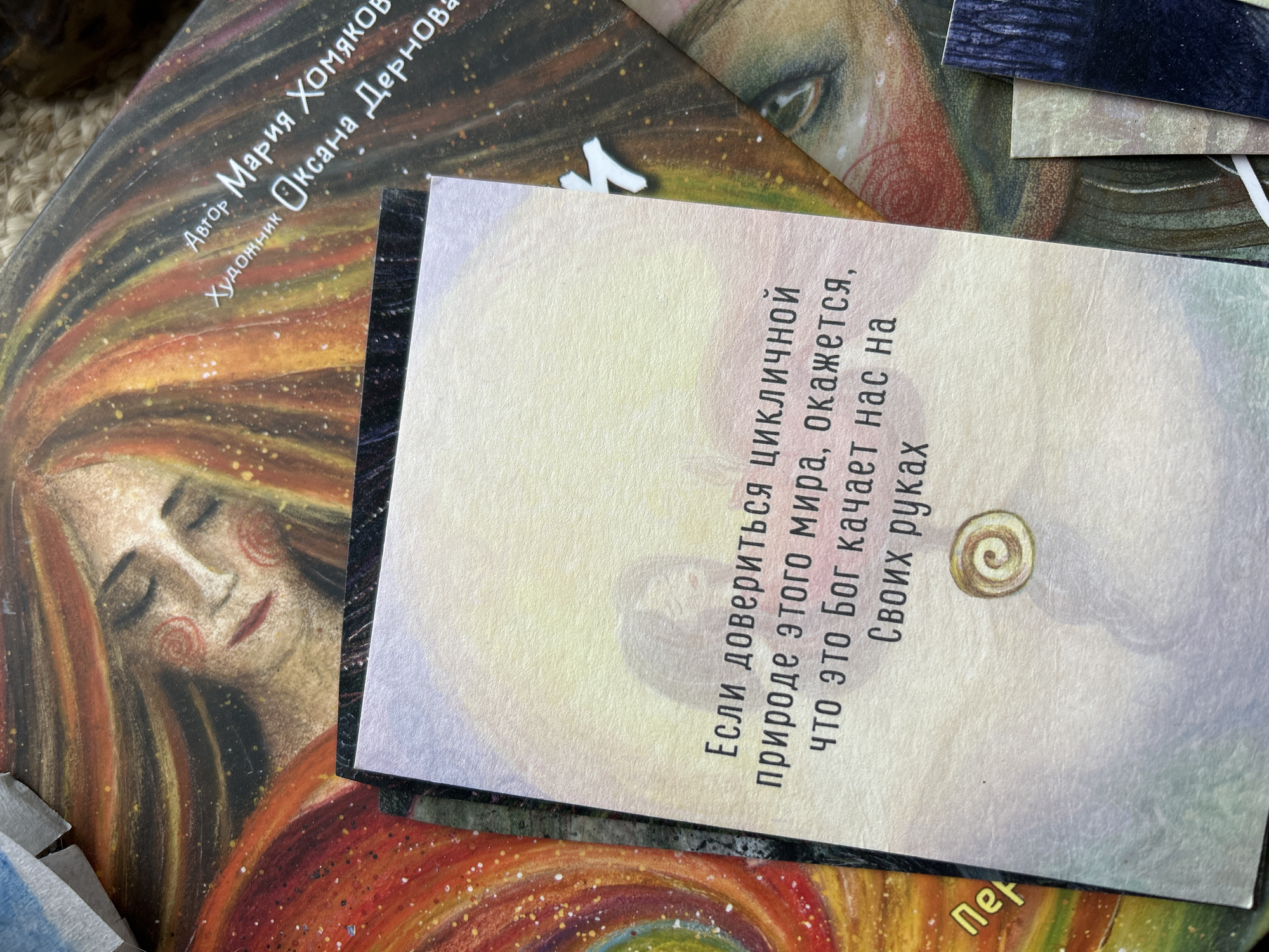 The image shows a close-up shot of several cards or book covers scattered on a surface. One card features a portrait of a woman with closed eyes and flowing hair, rendered in warm colors. Overlapping this card is another card with a light-colored background and black text in Cyrillic, along with a small, circular emblem. Part of another card with an eye is also visible. The text on the main card reads "Автор Мария Хомяков" and "Художник Оксана Дернова," indicating the author and artist. The other card reads, "Если довериться ЦИКЛИЧНОЙ природе этого мира, окажется, что это Бог качает нас на Своих руках", a philosophical quote.