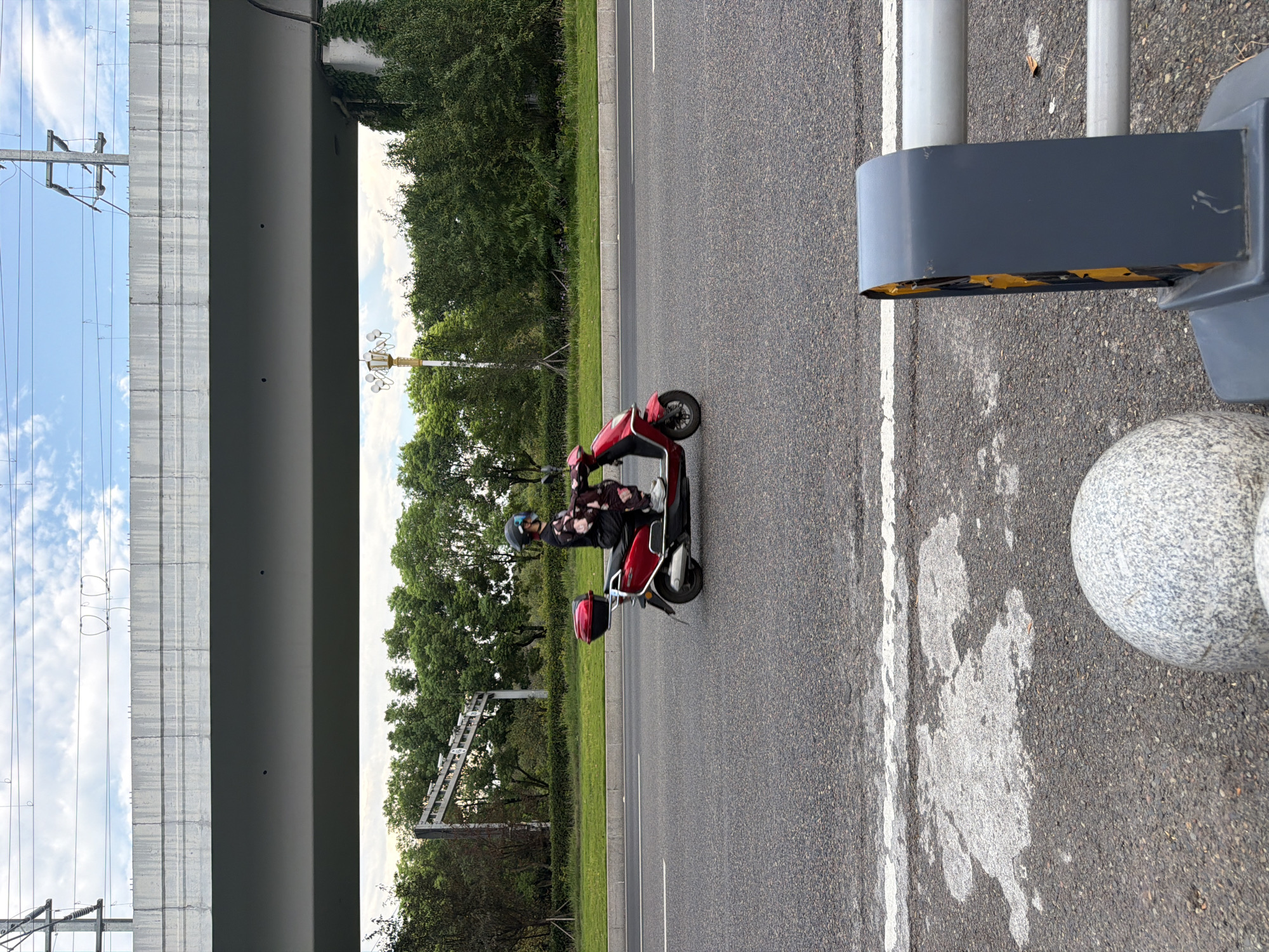 A person wearing a helmet and a patterned outfit is riding a red scooter on an asphalt road. The scooter has a black storage box on the back. The road is bordered by a green lawn and trees. Overhead, a concrete overpass is visible with power lines running above it. In the foreground, a grey, spherical bollard and a dark grey barrier with yellow accents are present. The sky is cloudy. The scene appears to be on an overpass or elevated roadway in Jiangjin, China.