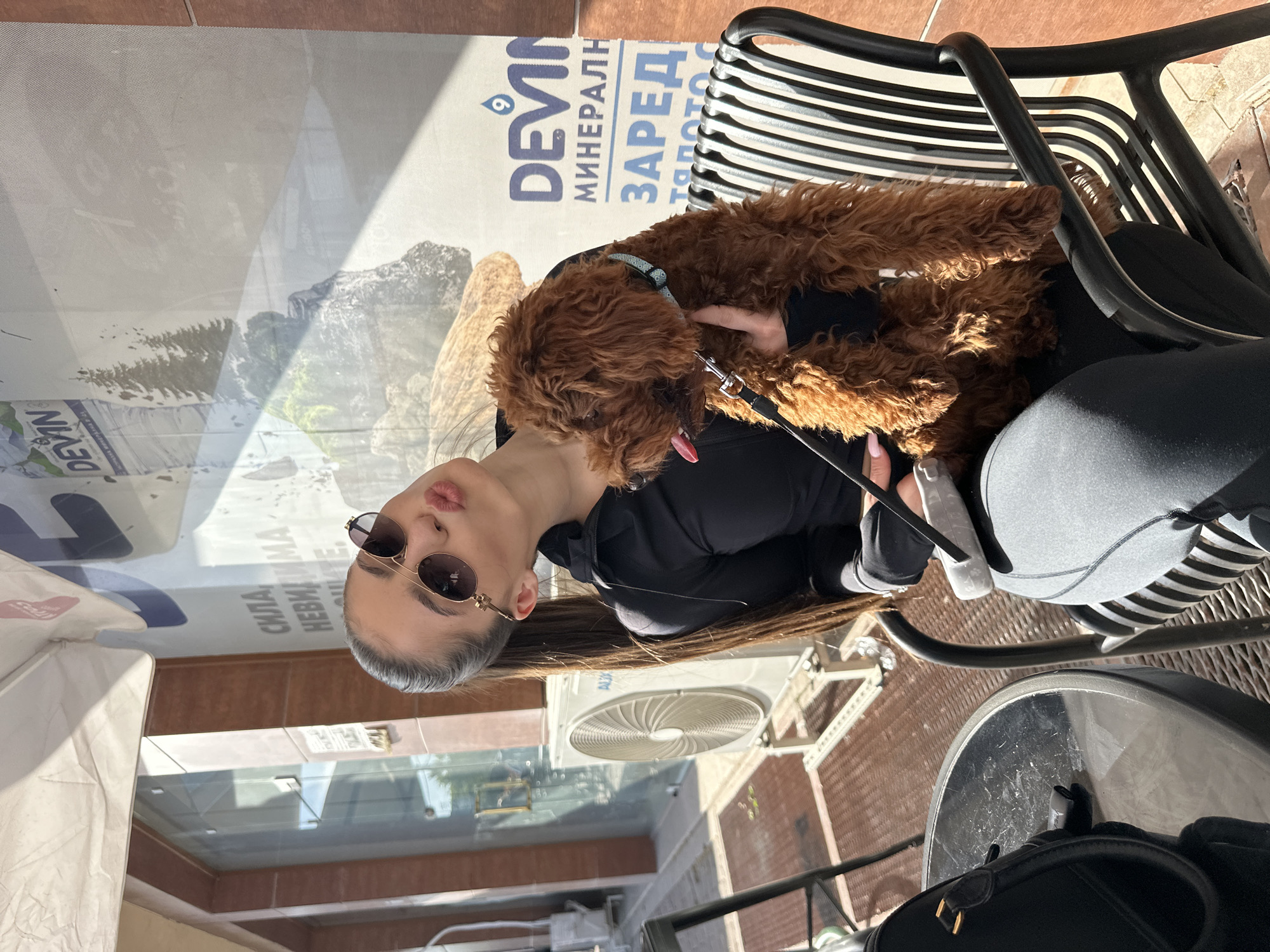 An outdoor scene in Plovdiv, Bulgaria, captured during a bright, sunny day, likely at a cafe or restaurant. The image features a young woman and a small, reddish-brown, fluffy dog.
The woman, with long dark hair, is wearing a black top and round sunglasses. She is seated, with her lips slightly puckered in a playful pose, holding the dog on her lap or arms. Her nails appear to be light-colored. The small dog, possibly a poodle or doodle, has thick curly fur and wears a light blue collar. Its tongue is slightly visible, and it appears relaxed and comfortable next to the woman.
They are seated on a black slatted chair, with a round, dark table visible in the foreground, partially obscured by a black bag. The background reveals architectural elements consistent with an urban setting. There are brown tiled surfaces, glass panels reflecting the surroundings, and an outdoor air conditioning unit mounted on a building wall.
A prominent feature in the background is a large advertisement banner or poster. Visible text on this banner includes "DEVIN" (a known Bulgarian mineral water brand, accompanied by a "9" symbol), "МИНЕРАЛНА" (Bulgarian for "mineral"), and "ЗАРЕД" (likely part of a word meaning "recharge" or "charge"). Other partially visible Bulgarian phrases like "СИЛА НА НЕВИДИМА" ("The power of invisible") and "НЕВЕЩИТЕ" ("things" or "elements") suggest a slogan related to natural power or vitality. The banner also displays imagery of mountains and water, aligning with the mineral water theme.
The bright sunlight casts distinct shadows, indicating clear weather. The overall impression is one of leisure and affection, as the woman poses with her pet in a casual, public setting.