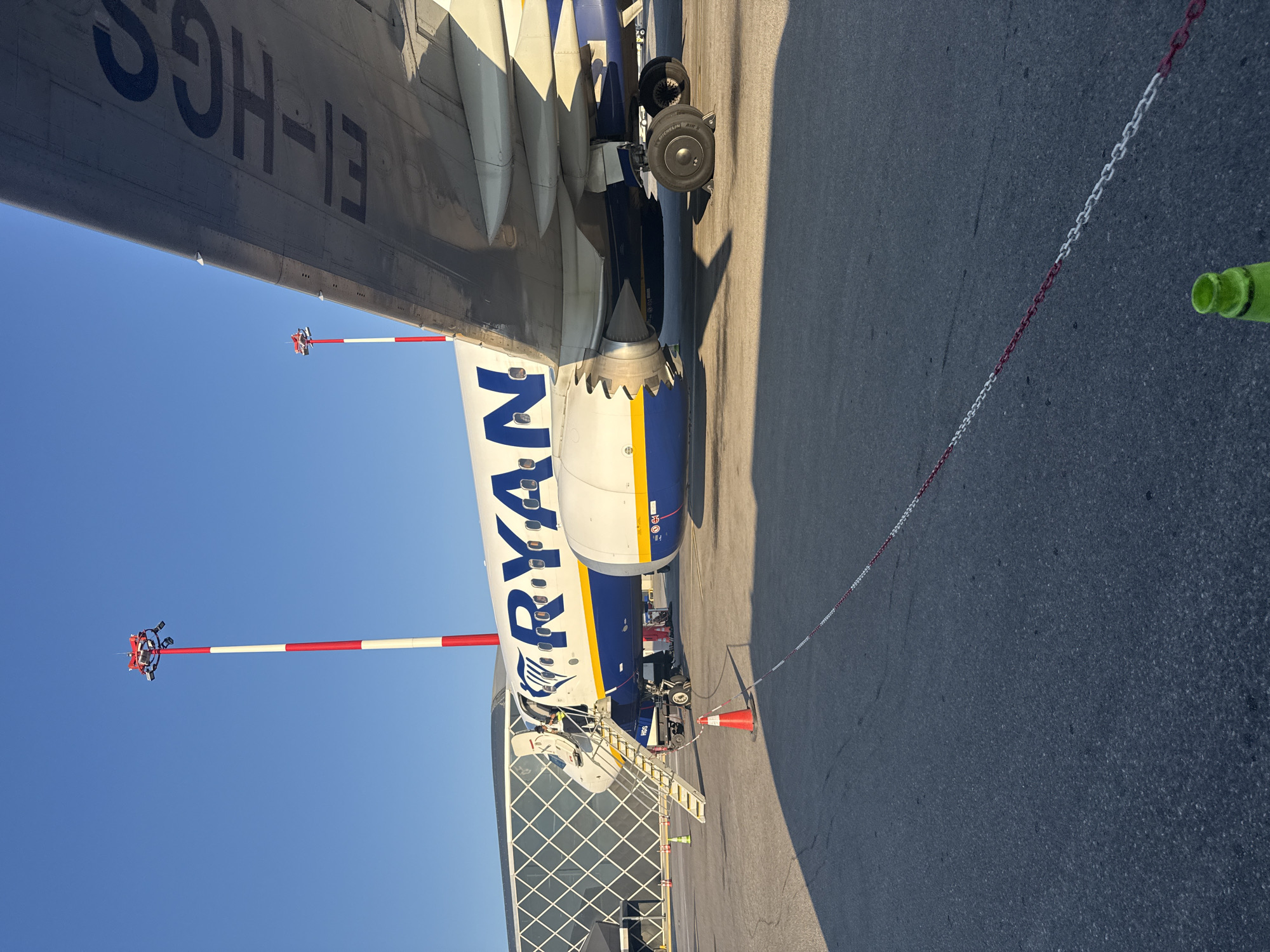 A Ryanair airplane is parked at an airport gate. The plane's registration number, EI-HGC, is visible on its wing. A set of mobile stairs is connected to the plane's forward cabin door, and a person is visible on the stairs. The airport terminal, a modern structure with a glass facade, is located behind the plane. Two tall poles with red and white stripes and lights are positioned near the plane. The ground is asphalt, with yellow and orange traffic cones and a red and white safety chain marking a boundary. The sky is clear and blue. The scene appears to be the Thessaloniki Airport in Thermi, Greece.