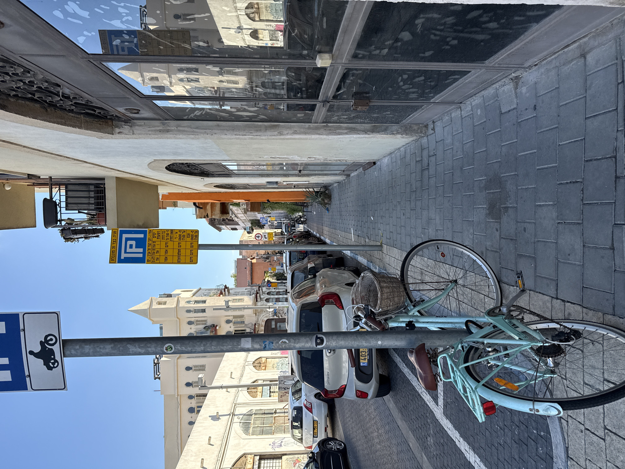 The image depicts a street scene in Tel Aviv, Israel. In the foreground, a light blue bicycle with a wicker basket on the handlebars is parked next to a metal pole. The bicycle has a brown leather seat and a metal rack on the back. The pole has two signs attached: a blue sign with a white outline of a motorcycle and a smaller white rectangle underneath, and a yellow sign with a blue "P" symbol and text in Hebrew. The street is paved with gray interlocking bricks, and white painted lines demarcate a parking area.
Several cars are parked along the street. A silver Kia Picanto is prominent in the mid-ground, with its rear facing the camera. Other vehicles, including a white Suzuki Swift, are visible further down the street. Buildings line the right side of the street, characterized by a mixture of architectural styles. The building closest to the viewer has a light-colored facade with large, reflective glass doors and windows. Further down the street, other buildings display cream-colored walls and arched windows.
The sky is clear and blue, indicating a sunny day. The overall environment suggests a typical urban street with parking regulations and pedestrian access. The presence of the bicycle and the parked cars indicates activity within the city. The signs suggest designated parking zones.
