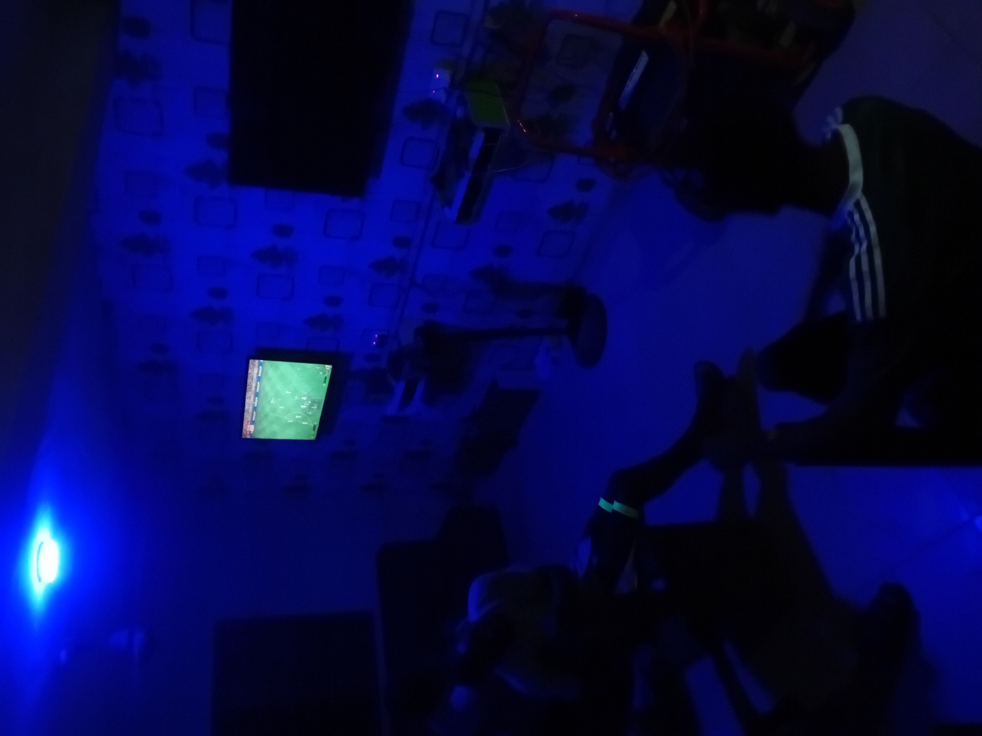 This image captures a dimly lit indoor scene dominated by a strong blue light. A bright, circular blue light source is prominently featured on the left wall, illuminating the room in cool tones. The walls, and possibly the ceiling, display a repeating pattern of squares and indistinct cloud-like shapes.
In the foreground on the right, a person is seen in profile, heavily silhouetted against the blue light. They are leaning forward, wearing a dark top with three distinct white stripes on the shoulder and sleeve, suggestive of an Adidas brand item. A glowing green band or bracelet is visible on their left wrist, shining brightly under the blue lighting. Further into the room, another person's silhouette can be vaguely discerned in the middle ground, adding to the sense of occupancy.
On the left wall, a small television screen is mounted, displaying what appears to be a sports game, likely soccer or football, given the visible green pitch. Above this screen and extending towards the upper center, various items are suspended, including a rectangular green object and several wires. A red structural frame, possibly part of a bed or a piece of furniture, is partially visible in the upper right portion of the image.
The scene conveys an evening or nighttime setting, with the artificial blue light creating a relaxed and focused atmosphere, perhaps centered around entertainment. No discernible text is visible on the screen or elsewhere in the image.