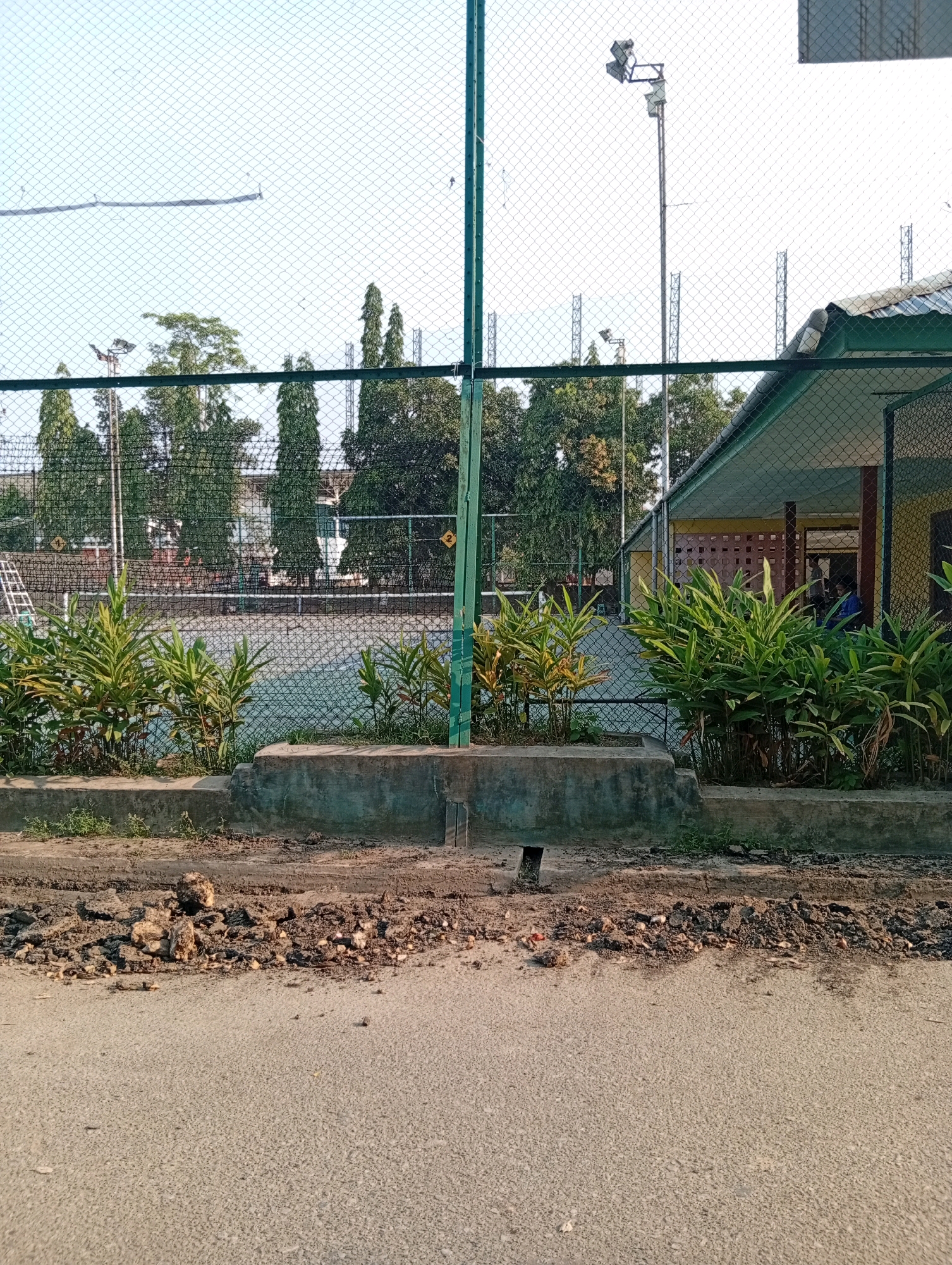 The image shows a tennis court with a chain-link fence in the foreground. Behind the fence, there is a tennis court with a light blue surface. There are trees and buildings in the background. The ground in front of the fence is made of asphalt and gravel. The scene appears to be a tennis court in an outdoor recreational area. The presence of tall, slender trees suggests a tropical or subtropical climate, consistent with Samarinda, Indonesia. There are no people visible in the image, and no specific actions or interactions are depicted. The environment is an outdoor sports facility.
