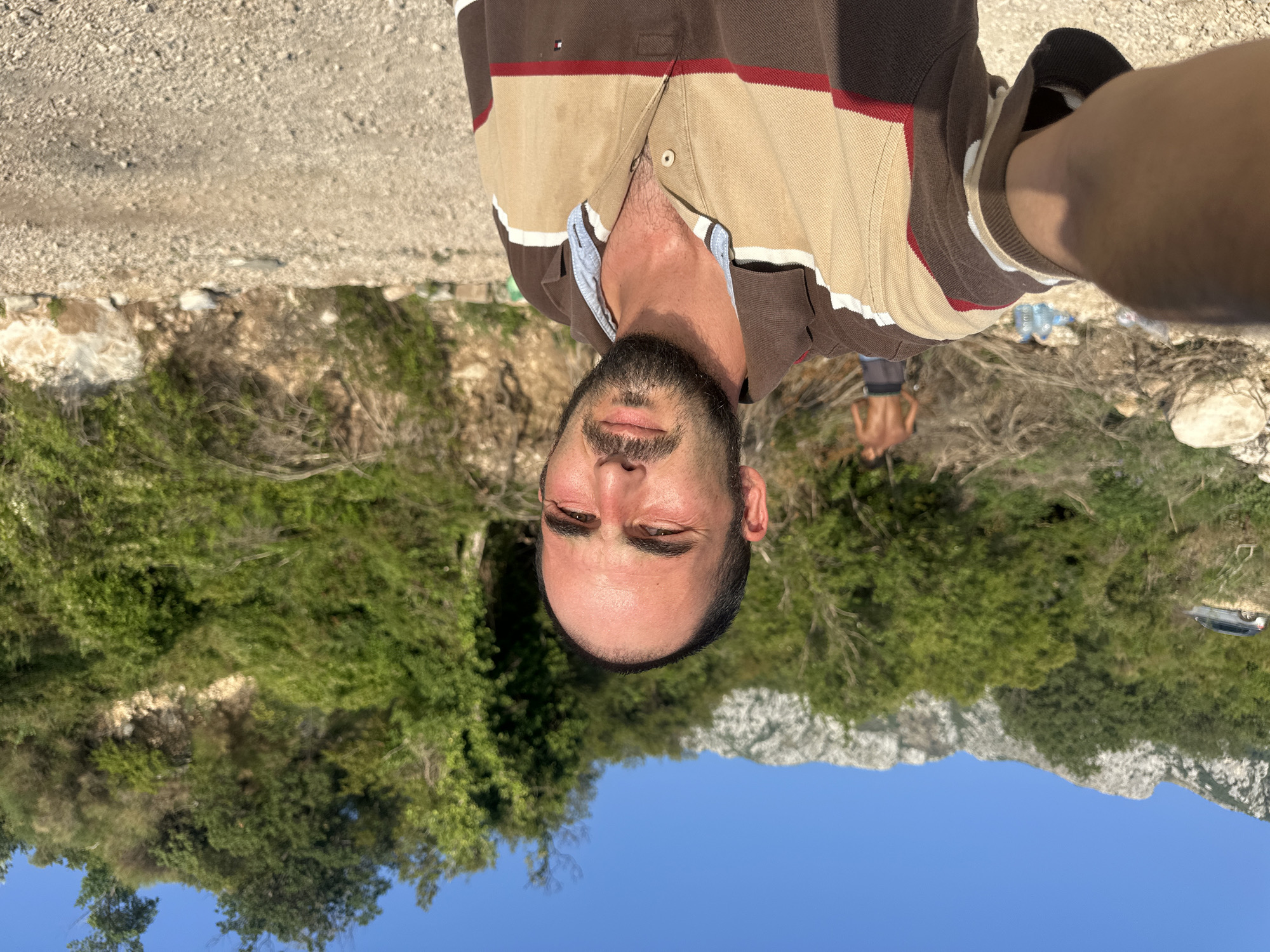In Sveti Stefan, Montenegro, a man with dark hair and a beard takes a selfie in an outdoor setting. He is wearing a brown, beige, and red striped polo shirt. In the background, a second shirtless man with dark hair stands near a pile of branches and some plastic water bottles. A silver SUV is parked on a dirt road in the distance, adjacent to a lush, green hillside with a rocky incline. The ground is covered in gravel, and the sky is a clear blue.