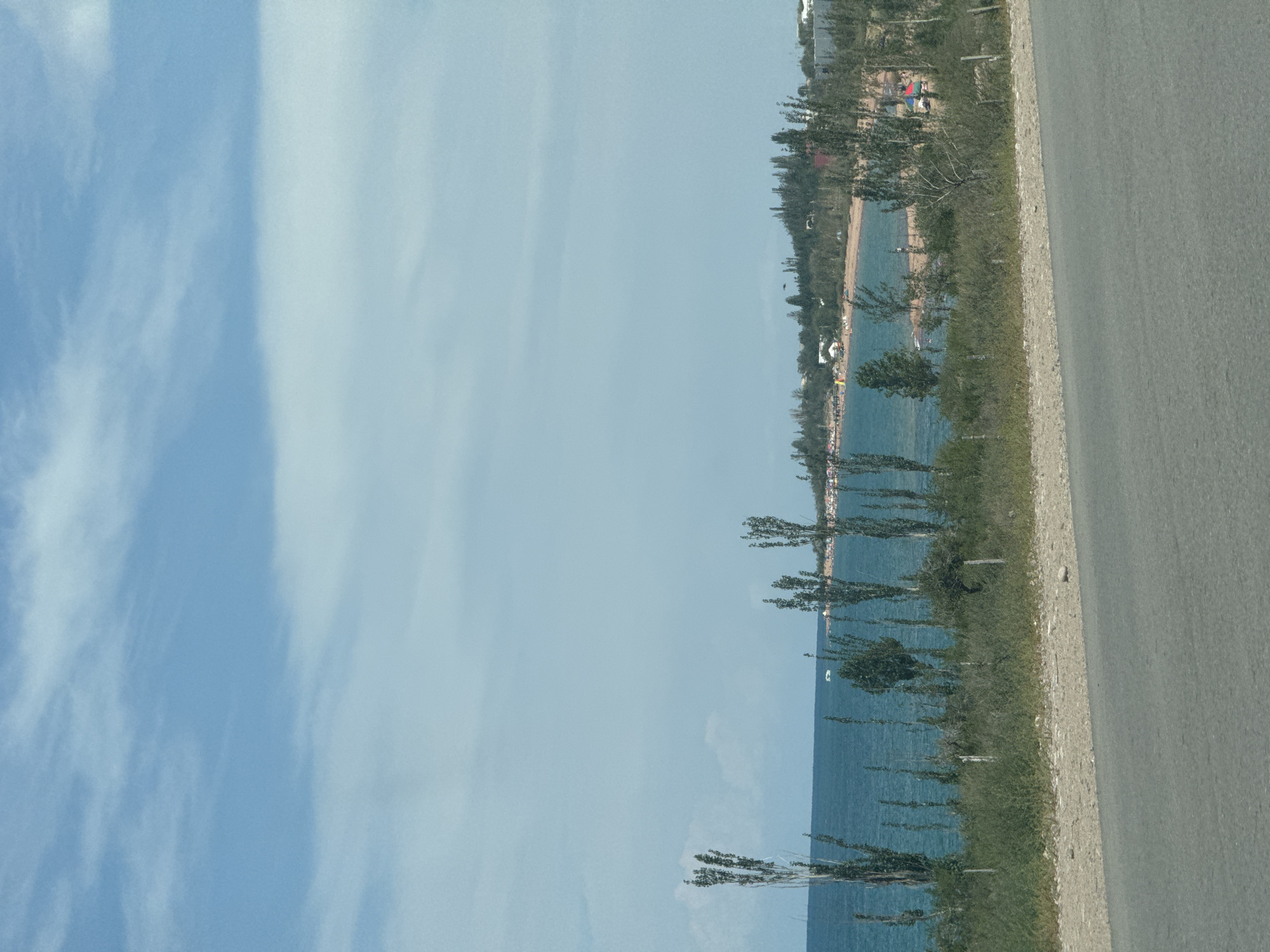 An asphalt road runs along the bottom of the image, separating the viewer from a grassy embankment. Beyond the embankment, a body of turquoise water stretches to the horizon. Tall, slender trees with sparse foliage dot the shoreline. In the distance, a sandy beach is visible, populated by a few people and colorful umbrellas. Buildings and more trees form a backdrop to the beach. The sky is a pale blue with scattered white clouds. The scene appears to be Kaji-Say, Kyrgyzstan, given the characteristic landscape and architecture.
