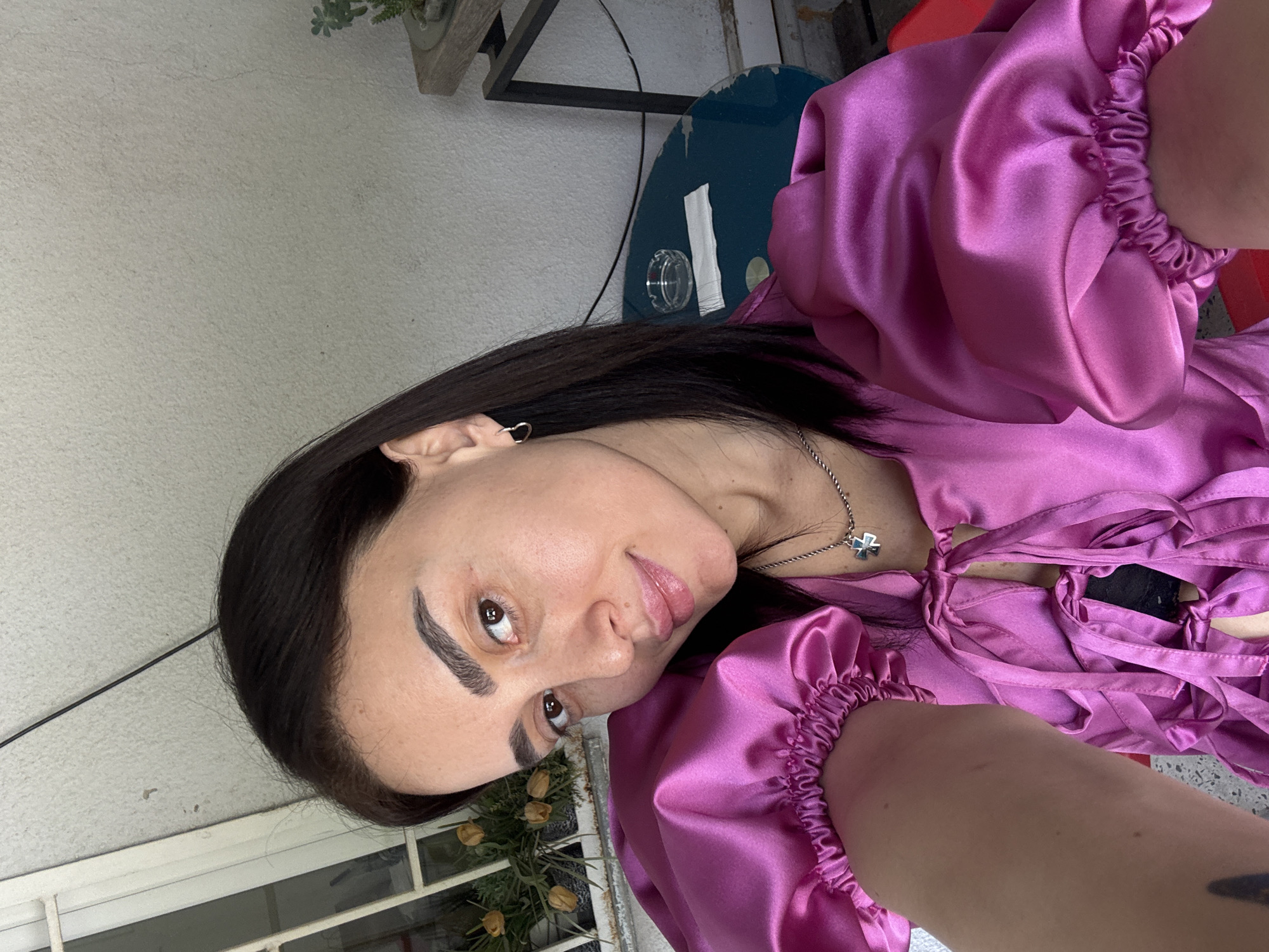 A woman is seen in a selfie-style photograph. She is wearing a bright pink satin blouse with ruffled sleeves and ties at the neckline, along with a silver necklace with a pendant. Her dark hair is styled smoothly to one side, exposing one ear with a small earring. The woman's makeup includes defined eyebrows and light lip color.
In the background, a white wall is visible, along with a window with a view of some plants. To the upper left, there is a glimpse of a metal table leg and a blue round table. The location appears to be an outdoor or semi-outdoor area, potentially in a residential setting. There is no specific evidence in the photograph to confirm the location is Tbilisi, Georgia.