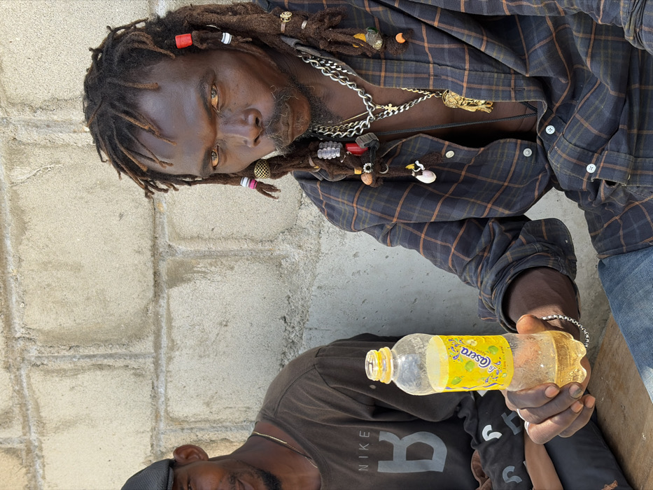 In Igboefon, Nigeria, two males are seated outdoors against a light gray, unpainted concrete block wall. The primary subject, a dark-skinned male with elaborate dreadlocks adorned with red, white, black, and brass beads and rings, occupies the right side of the frame. He wears an open dark blue and brown plaid long-sleeved shirt, revealing his bare chest, along with blue jeans, multiple silver-colored chain necklaces (one featuring a cross pendant, another a large gold-colored lion head pendant), and a silver-colored bracelet on his left wrist. He holds an open, clear plastic bottle, approximately half-filled with a yellow liquid and bearing a yellow "Osara" label depicting limes, in his left hand, angled towards the viewer. His gaze is direct and intense, with a visible mustache and goatee. To his left, a second dark-skinned male is partially visible, wearing a dark flat cap and a dark brown long-sleeved t-shirt with a "NIKE B" logo. A thin gold-colored chain is visible around his neck, and he appears to be smiling slightly.