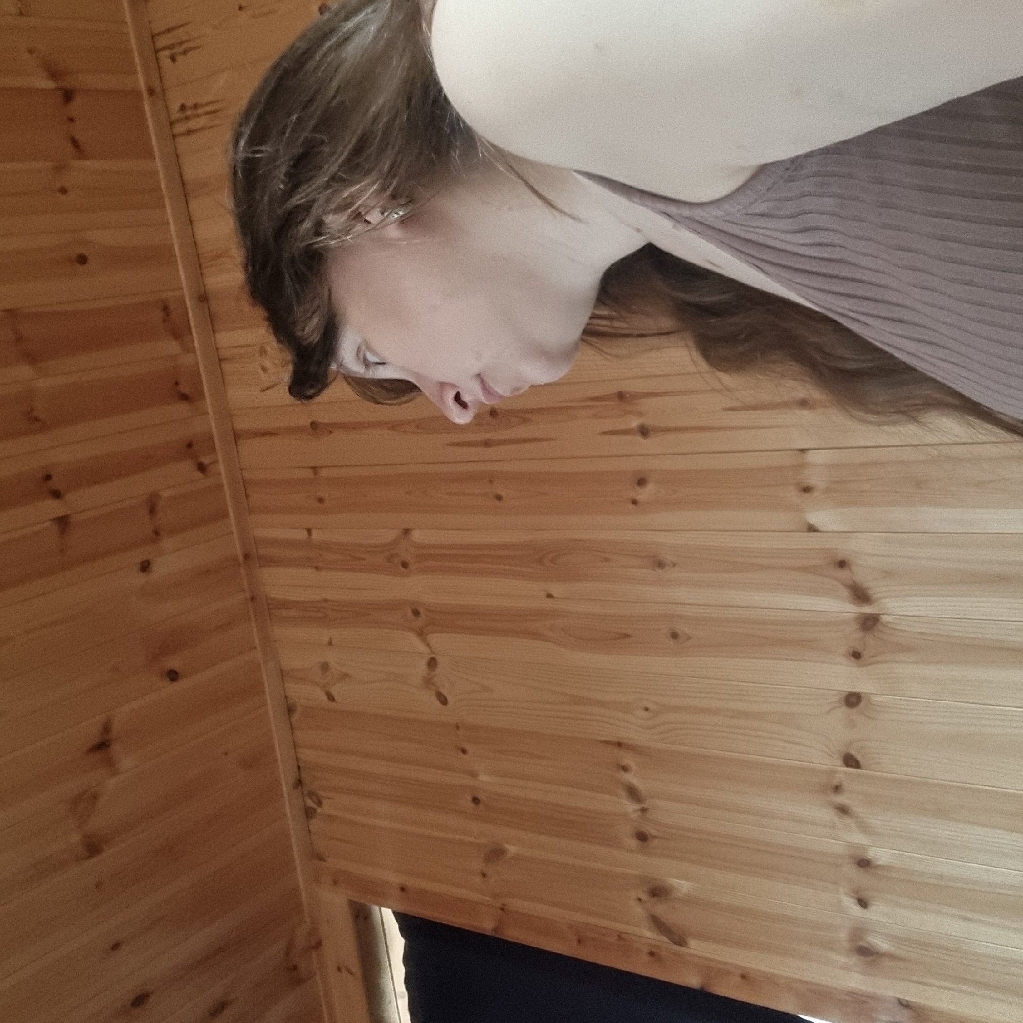 A medium shot shows a woman from the side in an indoor setting with wooden walls and ceiling. The woman has long, brown hair and is wearing a light purple, ribbed tank top. She is looking to her left, away from the camera. The background consists of vertically paneled light brown wood walls and a sloped wooden ceiling. A dark object is partially visible on the left side of the frame. The scene appears to be a room with natural wood paneling.