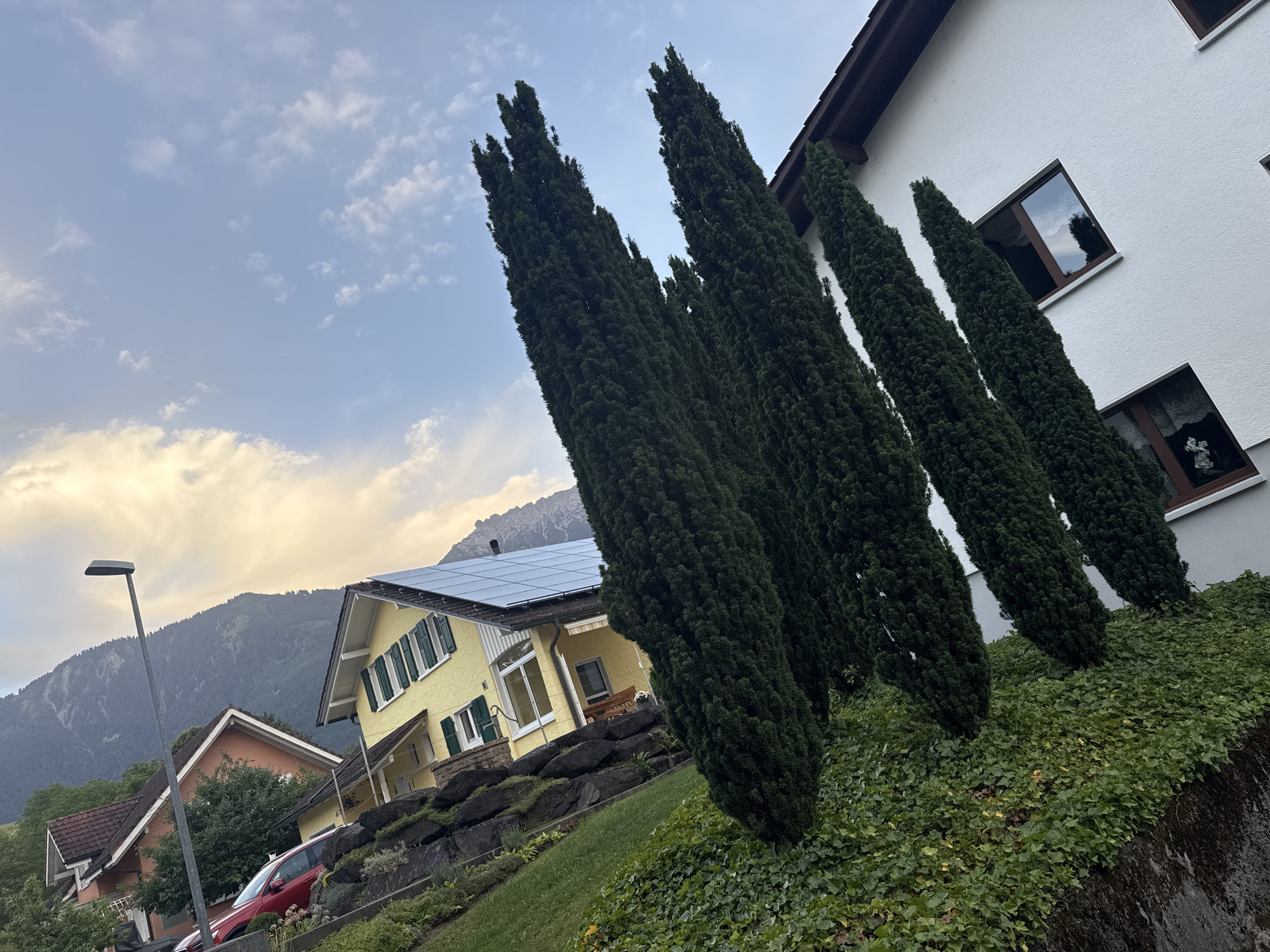 This is a wide-angle, eye-level outdoor photograph taken in Nendeln, Liechtenstein, featuring a residential area with houses and natural elements. The foreground is dominated by a steep, grassy slope covered in lush green ivy. Several tall, slender cypress trees stand prominently on the slope, their dark green foliage creating a strong vertical line. To the right, a portion of a white, modern house is visible, characterized by its angled roofline with dark trim and multiple windows with dark frames. The windows reflect the sky and surrounding trees.
In the middle ground, a bright yellow house with green shutters and a light-colored awning is situated on a lower level. This house has a roof covered with solar panels. To the left of the yellow house, a smaller, orange-roofed house is partially visible, nestled amongst trees. A red car is parked on a driveway in front of these houses.
The background consists of a mountain range covered in dark green trees, with some rocky, lighter-colored peaks visible at the top. The sky above is a pale blue, with scattered white clouds, some of which are tinged with yellow and orange, suggesting either early morning or late afternoon light. A modern, grey street lamp with a rectangular head stands on the left side of the frame, near the yellow house. The overall scene depicts a tranquil, well-maintained residential environment within a mountainous landscape.