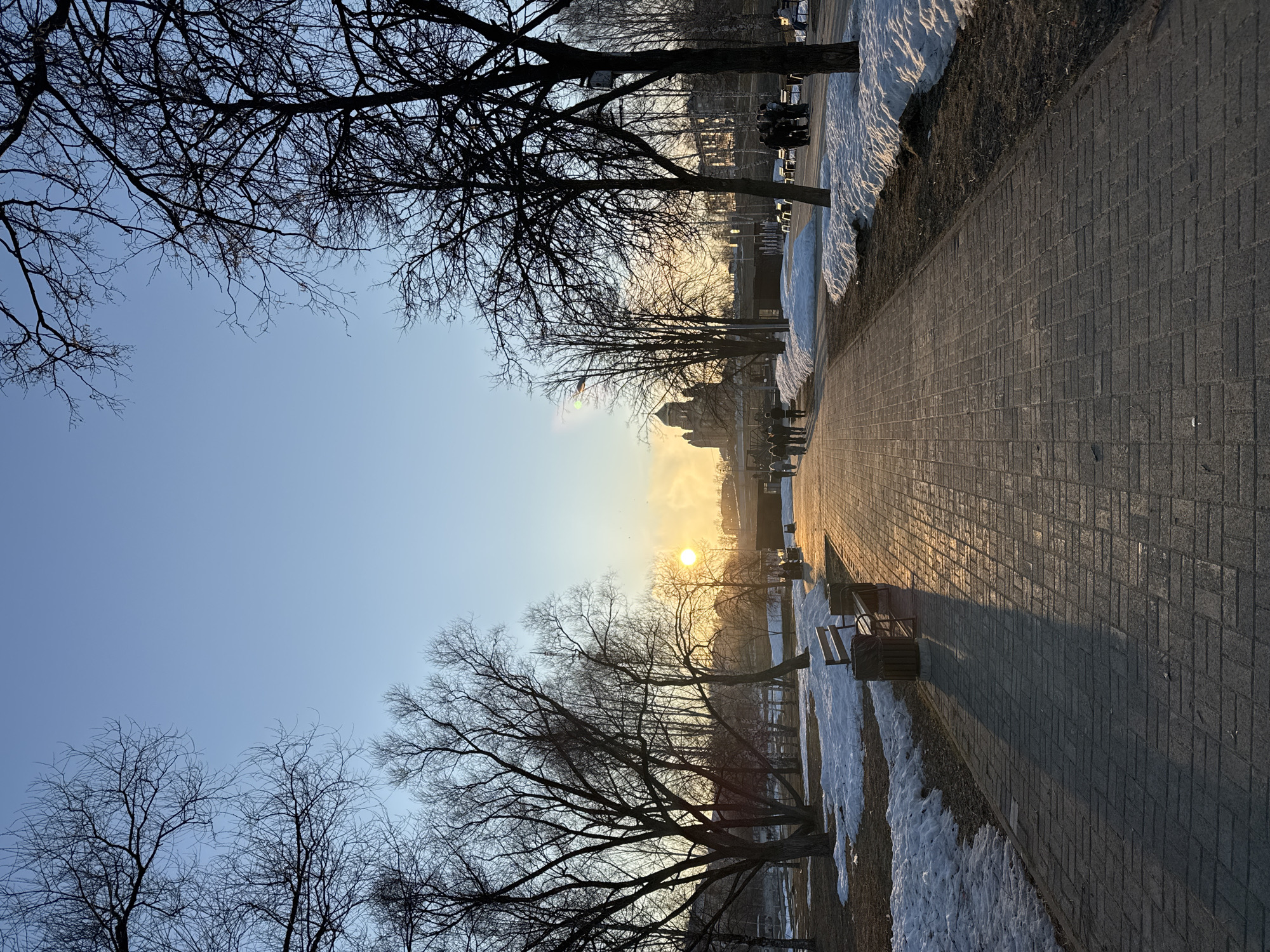 The image depicts a brick walkway in a park-like setting in Berezniki, Russia, during the late afternoon or early evening. The sun, low on the horizon, is visible between bare trees, casting long shadows along the walkway. Residual snow is present along the edge of the path. Bare trees line both sides of the path. A few benches and waste containers are positioned near the path. Silhouetted figures can be seen in the background, indicating the presence of people. The sky is clear.