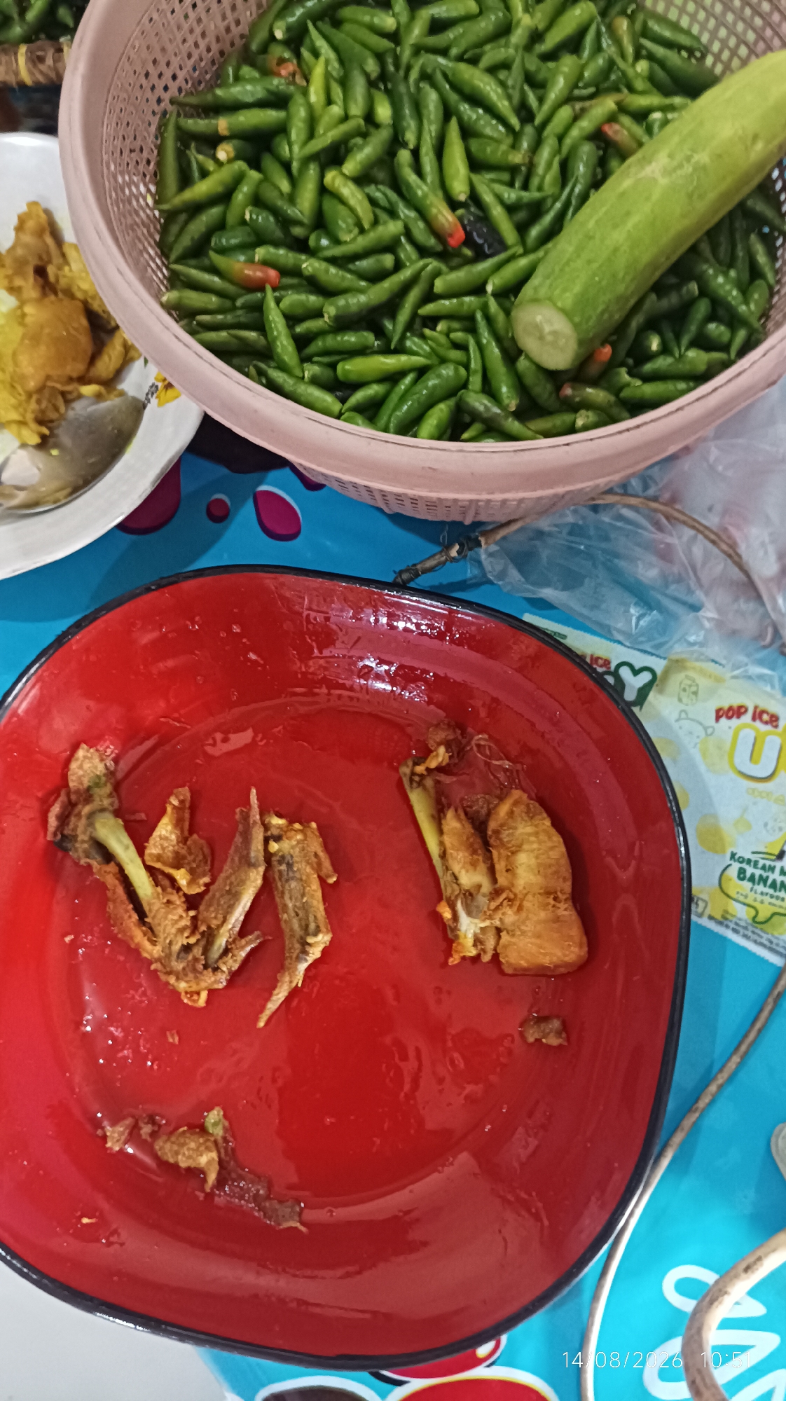 The image depicts a close-up, overhead view of a meal setting on a blue surface, possibly a table. In the upper portion of the frame, a light pink woven basket is filled with numerous small, green chili peppers, some with red or dark tips. A section of a cucumber, cut into a disk, rests on top of the chilies. To the left of the basket, a white plate holds a small portion of what appears to be fried food, possibly chicken, along with a spoon.
The lower part of the image focuses on a bright red, shallow bowl or plate. This red surface contains remnants of fried food, specifically small pieces of what look like bone and meat, indicating that food has been consumed from this dish. The red dish is slightly wet, with some scattered droplets.
In the background, to the right of the chili basket, a portion of a plastic wrapper with colorful text and imagery is visible. Some of the text appears to be in English, with words like "POP ICE" and "BANAN" discernible. A corner of the wrapper shows a logo that includes the letter "U".
The lower right corner of the image displays a timestamp: "14/08/2026 10:51".
The scene appears to be an informal meal, likely at a home or a casual eatery. The overall impression is one of fresh ingredients (chilies, cucumber) and a partially eaten meal. The presence of the timestamp suggests the image was taken on a specific date and time, possibly for personal record-keeping or to document the meal.
While the image itself does not provide explicit geographical markers, the presence of the "POP ICE" brand, which is popular in Indonesia, and the style of food preparation (fried items, fresh chilies) are consistent with culinary practices found in Indonesia. The city of Tasikmalaya, located in West Java, Indonesia, is known for its food culture, including the common use of chilies in its cuisine. Therefore, it is plausible that this scene is from Tasikmalaya, Indonesia, though without further contextual information, this remains an inference.
