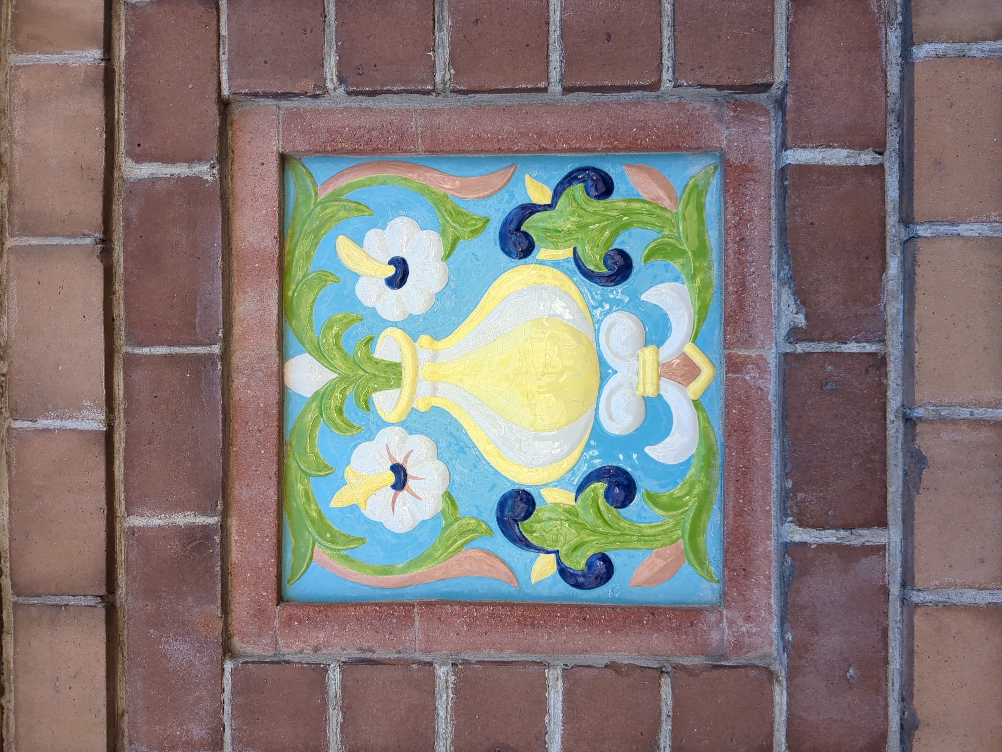 A square decorative ceramic tile is embedded within a brick wall, forming the central subject of the image. The tile features an ornate, symmetrical design on a light blue background. At the heart of the design is a stylized vase or jug, predominantly yellow with white accents, displaying a subtle crackled glaze. Surrounding this central element are vibrant green leafy scrollwork, white flowers with yellow petals and dark blue centers, and additional dark blue decorative flourishes.
The tile is framed by a border of reddish-brown bricks, which seamlessly integrate with the larger brick wall. The wall itself is constructed from bricks of varying reddish-brown and lighter tan hues, with distinct mortar lines visible between them, indicating some age and weathering. This architectural detail appears to be an outdoor embellishment. No human activity or specific event is taking place; the image focuses solely on this static decorative element. The lighting suggests it is daytime. There is no visible text. The location context for this architectural feature is Saint Petersburg, Russia.