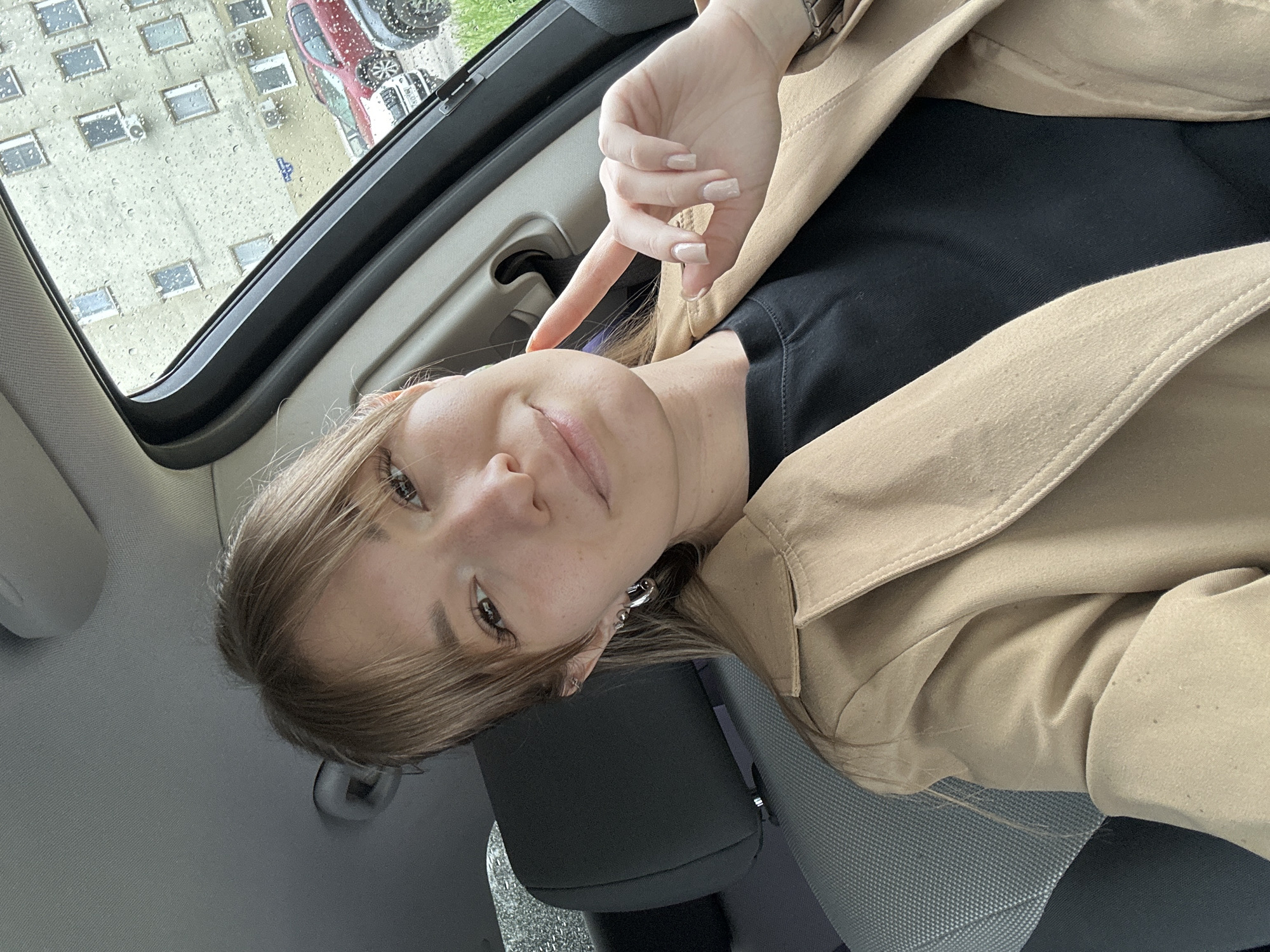 A woman with light brown hair, wearing a black shirt and a tan trench coat, is in a car. She is pointing to her face. The car appears to have a sunroof, through which an apartment building can be seen. Rain is visible on the sunroof glass. The apartment building has multiple windows. A red car and a white car are visible outside. Based on the architecture of the apartment building, the scene could be located in the city of Chaikovsky, Russia. The woman is sitting in the driver's seat.