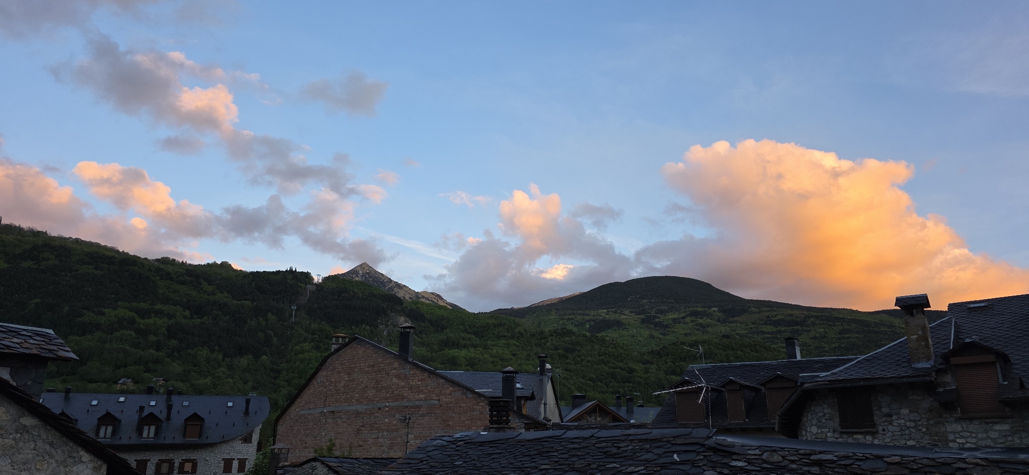 The image shows a view over the rooftops of buildings in the city of Benasque, Spain, towards the Pyrenees mountains. The roofs of the buildings are covered with dark slate tiles. In the background, two mountains covered in green trees are seen. A third mountain, with a sharp peak, is seen behind the others. The sky is a light blue color, with clouds tinged with pink and orange hues, indicating sunset or sunrise. A ski lift is visible on the side of one of the mountains. The architecture of the buildings is typical of the region, with chimneys protruding from the roofs.