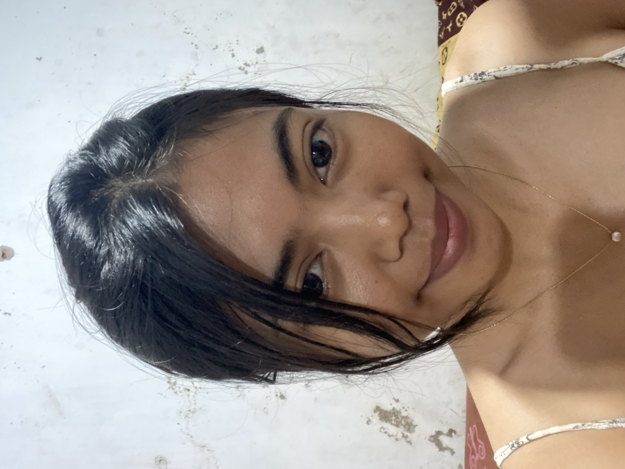 A young woman with dark hair is shown from the chest up, looking at the camera. She is wearing a white strap top with a subtle pattern. A delicate gold necklace with a single pearl pendant is visible around her neck. The background is a textured, off-white wall with some dark splotches and marks. The lighting appears to be indoor. The location is Madiun, Indonesia.