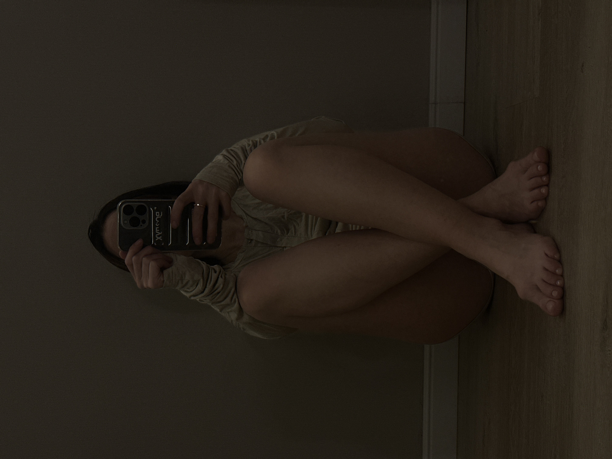 A woman is seated on the floor against a wall, knees bent and crossed in front of her chest. She is holding a smartphone up in front of her face, presumably taking a selfie. The woman is wearing a light-colored, long-sleeved shirt. The background consists of a dark wall and a lighter-colored floor with a seam line. It is not possible to determine the location within the city of Votkinsk, Russia from the image.