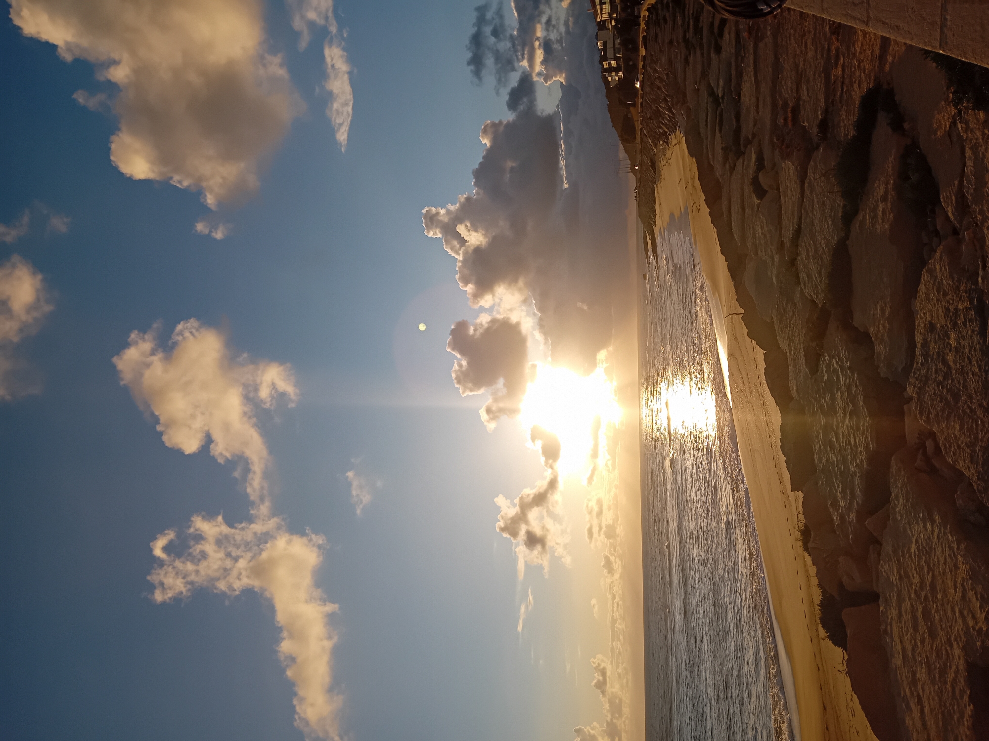 The image captures a seaside view at sunset. The sky is a mix of blue and golden hues, dotted with fluffy clouds. The sun, partially obscured by a large cloud formation, casts a bright glare, reflecting intensely on the water's surface. The ocean stretches out, with gentle waves visible near the shore. A sandy beach separates the ocean from a rugged, rocky cliff. On top of the cliff, the outlines of buildings suggest an urban area. The location is unknown, as the prompt specifies the city of Unknown, Unknown.