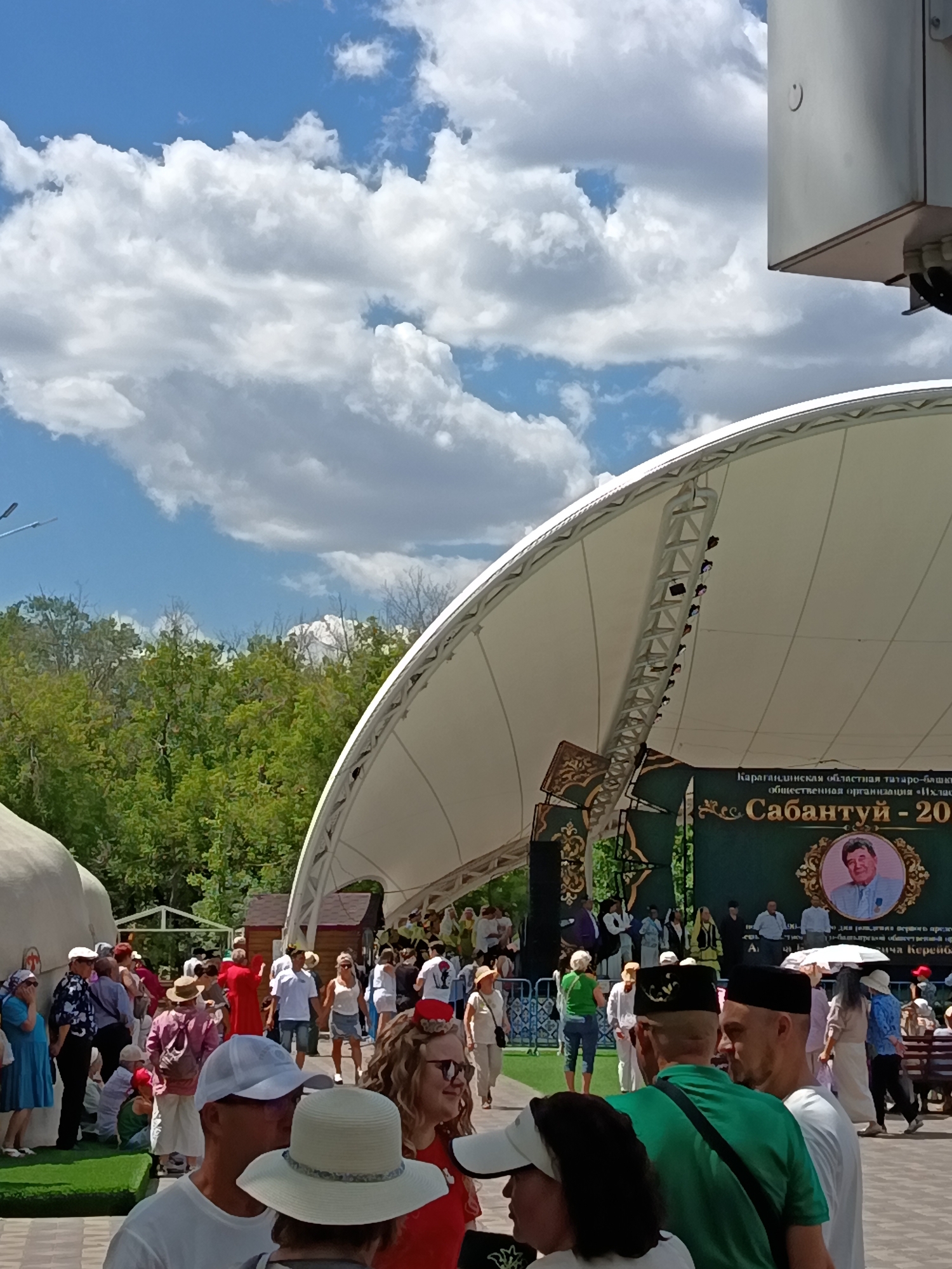 The image depicts an outdoor event on a sunny day with scattered clouds. A large, white, curved canopy structure dominates the upper right portion of the frame, supported by a metal truss system. Below the canopy, a green banner is displayed, featuring text in Cyrillic script and a portrait of a man in a framed oval. The banner reads: "Карагандинская областная татаро-башкирская общественная организация «Ильяс» Сабантуй - 20" (Karaganda Regional Tatar-Bashkir Public Organization "Ilyas" Sabantuy - 20).
A crowd of people is gathered in front of and around the stage area. Many are standing and appear to be observing the event. Several individuals are wearing traditional headwear, including black skullcaps and white hats. The attire of the attendees varies, with some in casual summer clothing and others in more formal or traditional garments. Some people are walking, while others are clustered together.
To the left of the stage area, there are trees and a low-lying building structure. A patch of green astroturf is visible in the foreground, with people sitting on it. The ground is paved. The overall atmosphere suggests a cultural festival or public gathering. The location is identified as the city administration of Qarağandı, Kazakhstan, implied by the presence of the banner.