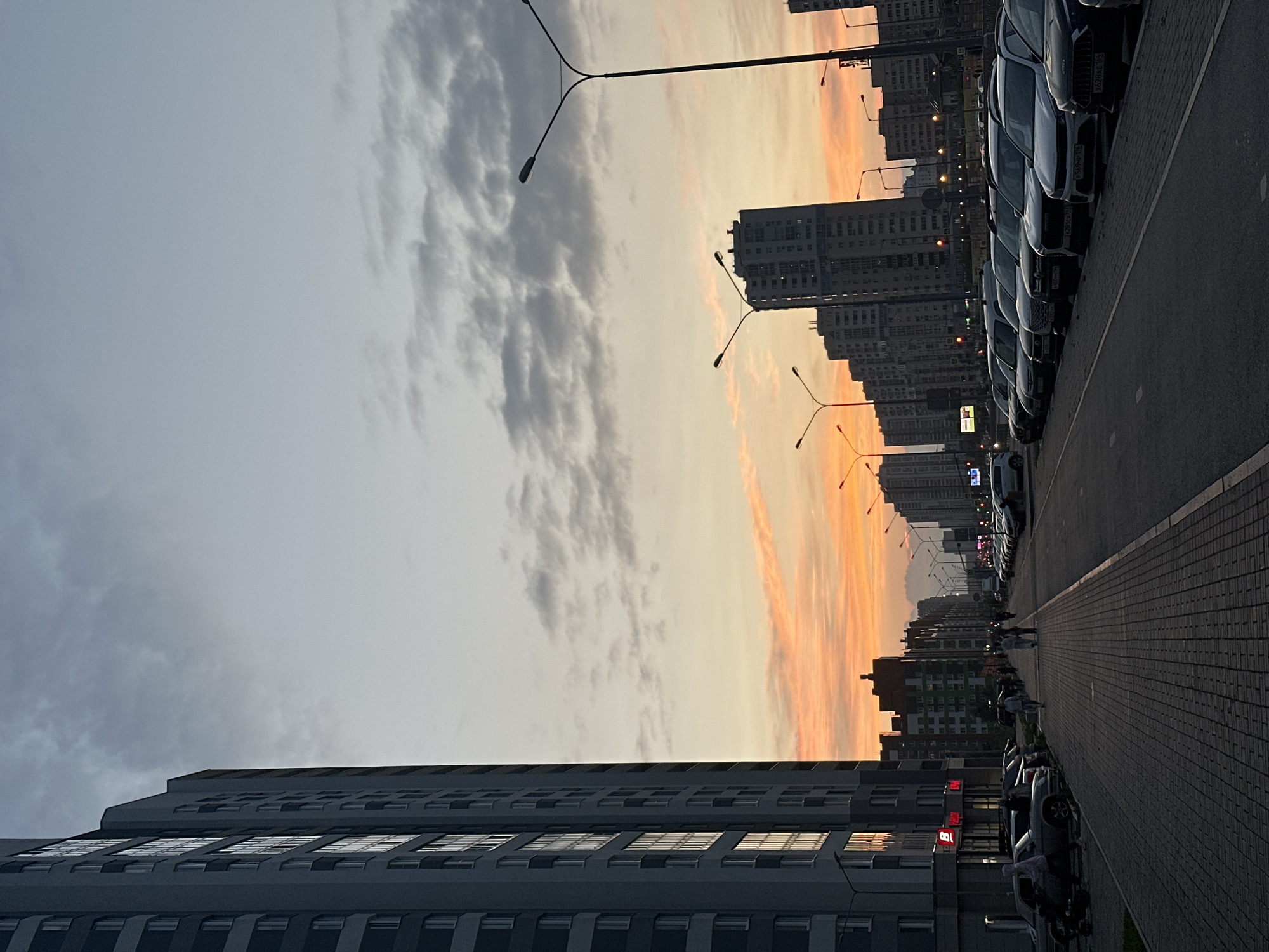 The image depicts a city street scene in Yekaterinburg, Russia, at dusk. A tall, modern residential building with numerous windows dominates the left side of the frame. The sky is a gradient of pale blue and grey with streaks of orange and pink from the setting sun.
A row of parked cars lines the right side of the street. The cars are of various makes and models, and some have visible license plates. A paved pedestrian walkway runs alongside the parked cars and in front of the residential building.
Several people are walking on the pedestrian walkway. One person is riding a scooter on the sidewalk near the left edge of the frame. Streetlights are visible along the street, some illuminated, and others not. In the background, more multi-story buildings stretch along the street, forming an urban landscape. The overall atmosphere is that of a quiet evening in a residential area of the city.