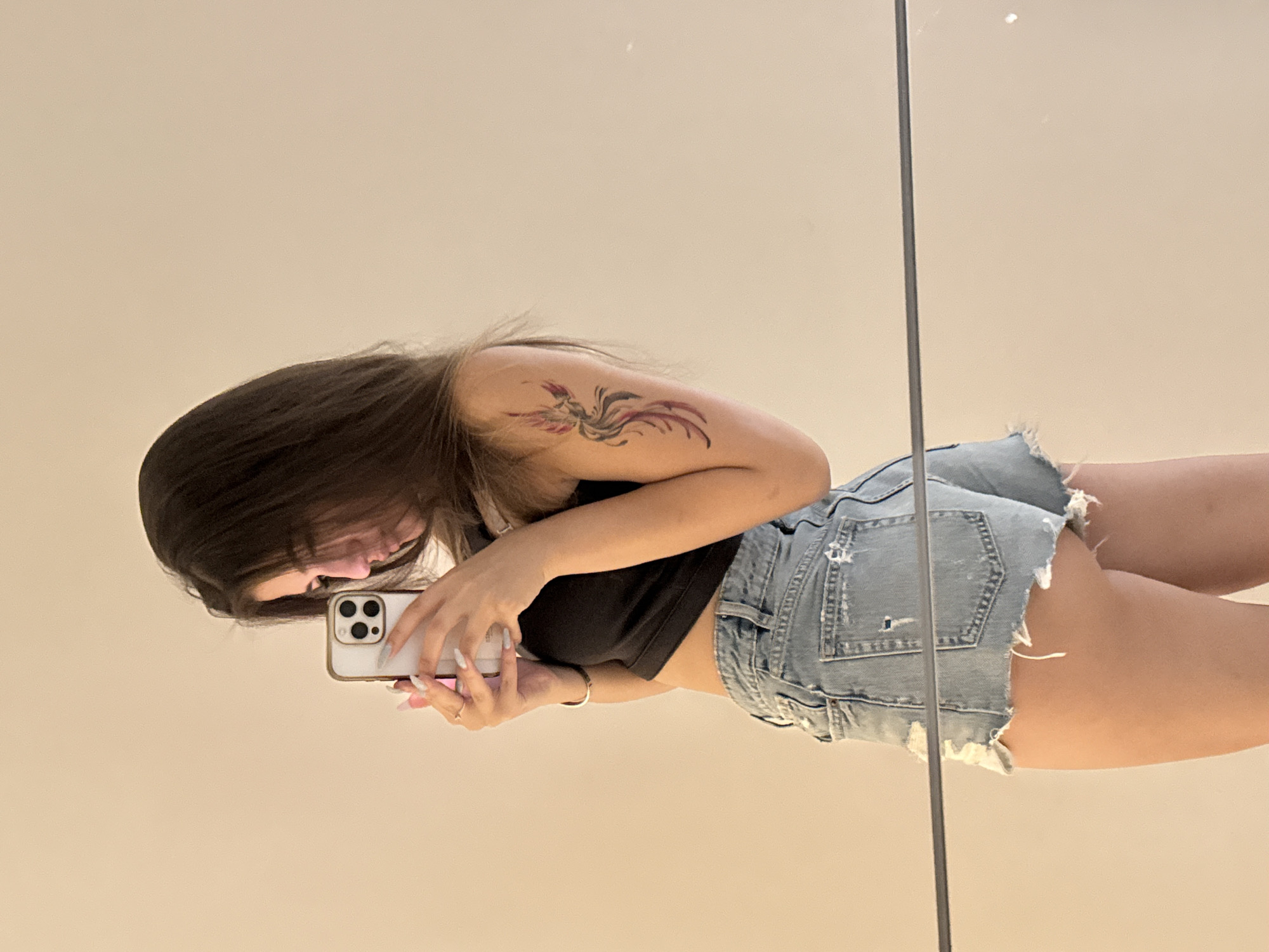 A young woman with long, dark hair is taking a mirror selfie. She is wearing a black crop top and denim shorts. A tattoo of a bird or phoenix is visible on her upper left arm. The background appears to be a plain wall, suggesting an indoor setting. No specific location in Sofia, Bulgaria, can be determined from the image.