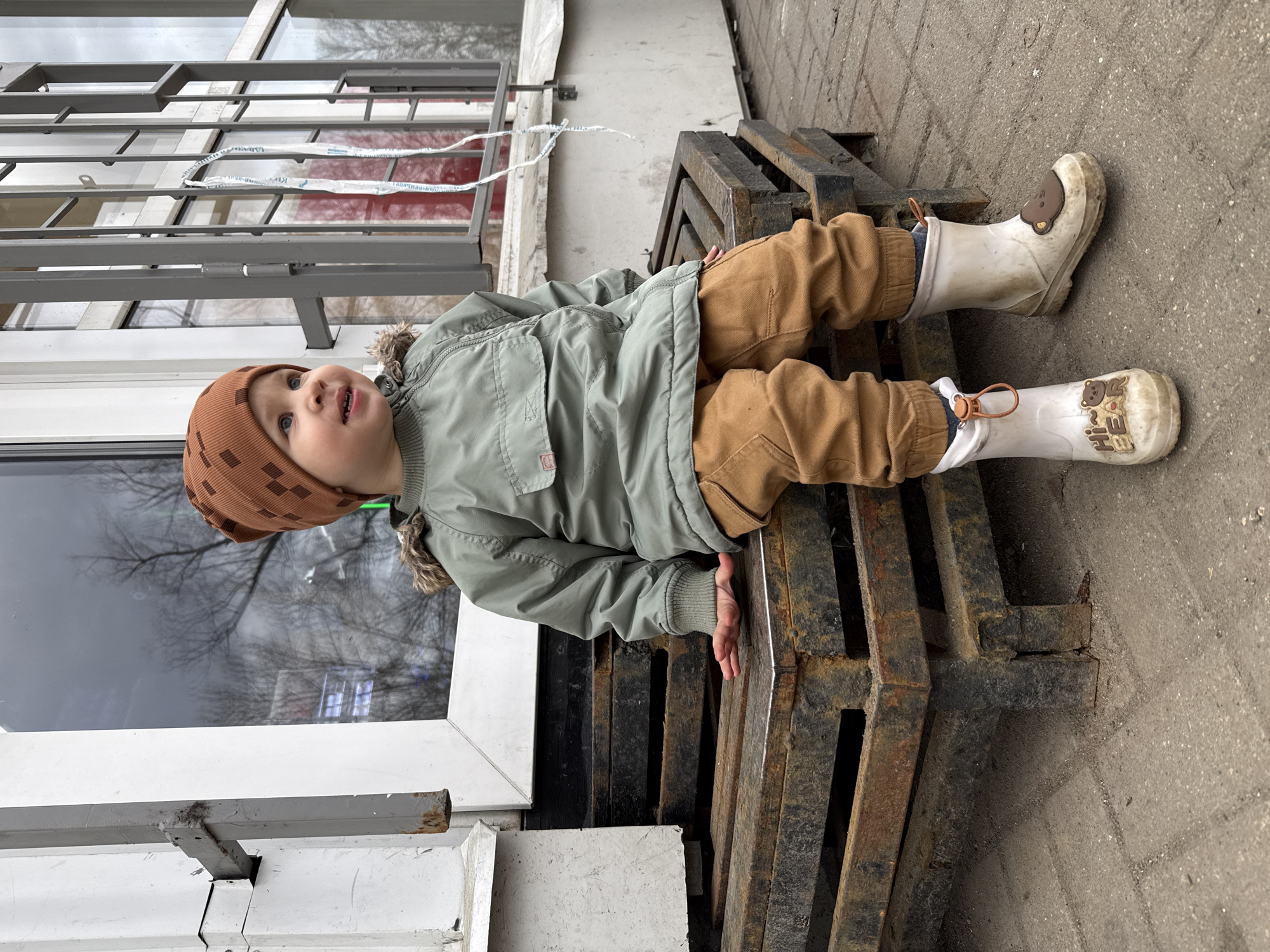 A toddler wearing a brown patterned beanie, olive green jacket with fur trim, brown pants, and white rain boots lies on a metal pallet. The pallet is resting on a concrete surface next to a white building with windows. The setting appears to be outdoors, possibly in Rusanovo, Russia.