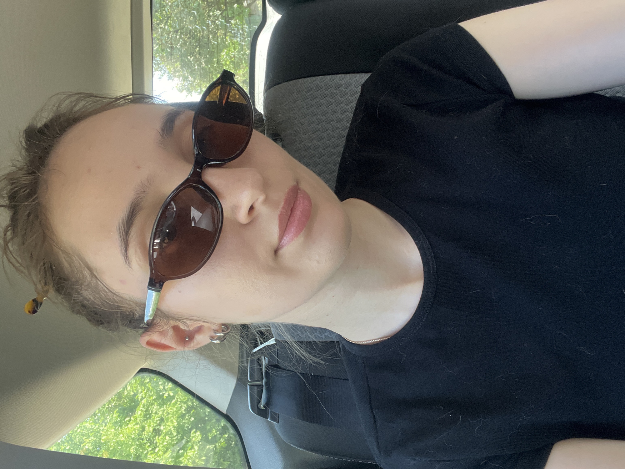 A young woman with blonde hair, wearing sunglasses and a black t-shirt, is shown from the chest up. She is indoors, possibly in a car, with a seatbelt visible across her chest. Her sunglasses are dark brown with tortoise-shell accents. She has earrings in both ears, including hoops on the left and a stud on the right. Her hair is pulled back and appears to have a tortoiseshell clip on the left side. The lighting suggests it is daytime, and the background visible through a window to the left indicates an outdoor setting with trees and sunlight. The woman's expression is neutral. The image is a selfie, taken from a slightly tilted angle. The t-shirt has a few visible bits of lint or pet hair on it.
