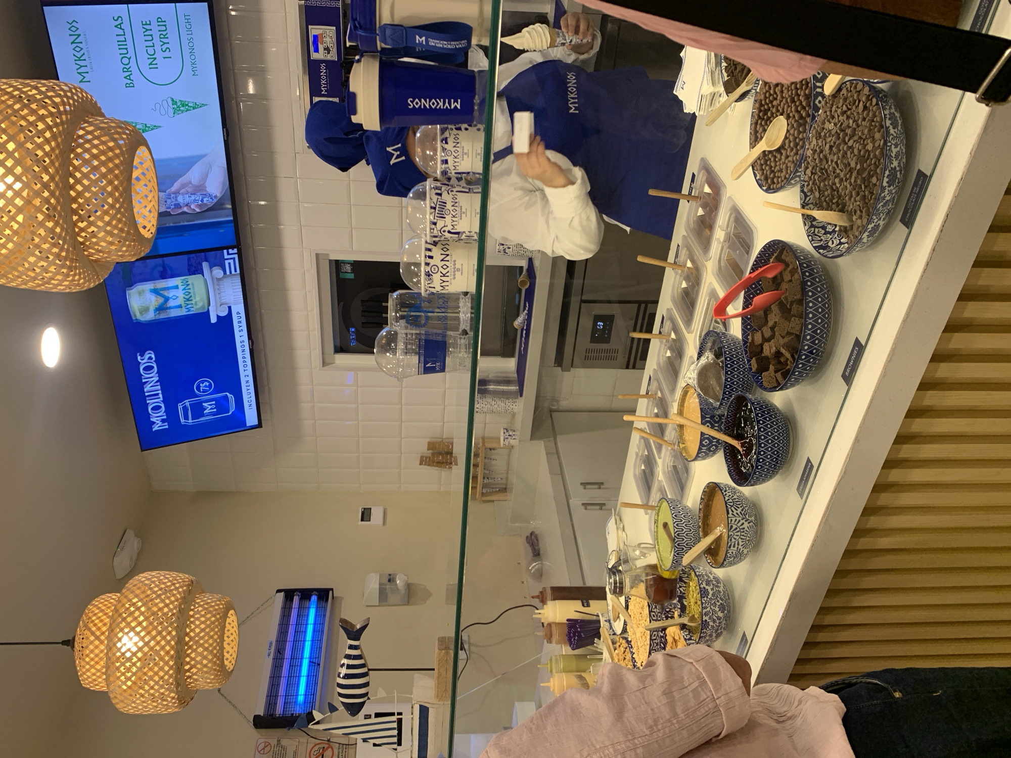 This image captures the interior of a frozen yogurt shop, likely "Mykonos Greek Frozen Yogurt," during operating hours. The scene is dominated by a long counter where desserts are prepared and displayed.
In the background, an employee wearing a white shirt, blue apron with "MYKONOS" branding, and a blue hat, is actively preparing an item, holding a cup and what appears to be a dispensing nozzle or an item to be added. Behind them are blue and white frozen yogurt machines. In the foreground, part of a customer in a pink top and blue jeans is visible, looking towards the counter.
The counter itself is a bright white surface showcasing numerous toppings in an organized manner. Various blue and white patterned bowls hold different items such as chocolate chips, brownie pieces, and other granular toppings. Clear containers also hold sauces and other sweets, accompanied by small wooden spoons or tongs for serving.
Above the counter, a large TV screen displays advertisements for "MYKONOS GREEK FROZEN YOGURT," showing product images and text like "BARQUILLAS INCLUYE 1 SYRUP" (waffles include 1 syrup) and "MYKONOS LIGHT." Another part of the screen mentions "MOLINOS" and "INCLUYEN 2 TOPPINGS 1 SYRUP."
The shop's decor includes two large, intricately woven wicker light fixtures hanging from the ceiling, casting a warm glow. On a wall to the left, a blue-light insect zapper is visible, next to a decorative blue and white striped wooden fish and what appears to be a nautical-themed decoration. The overall ambiance is clean, modern, and well-lit. The repetition of "MYKONOS" branding suggests a consistent theme throughout the establishment, located in Pampatar, Venezuela.