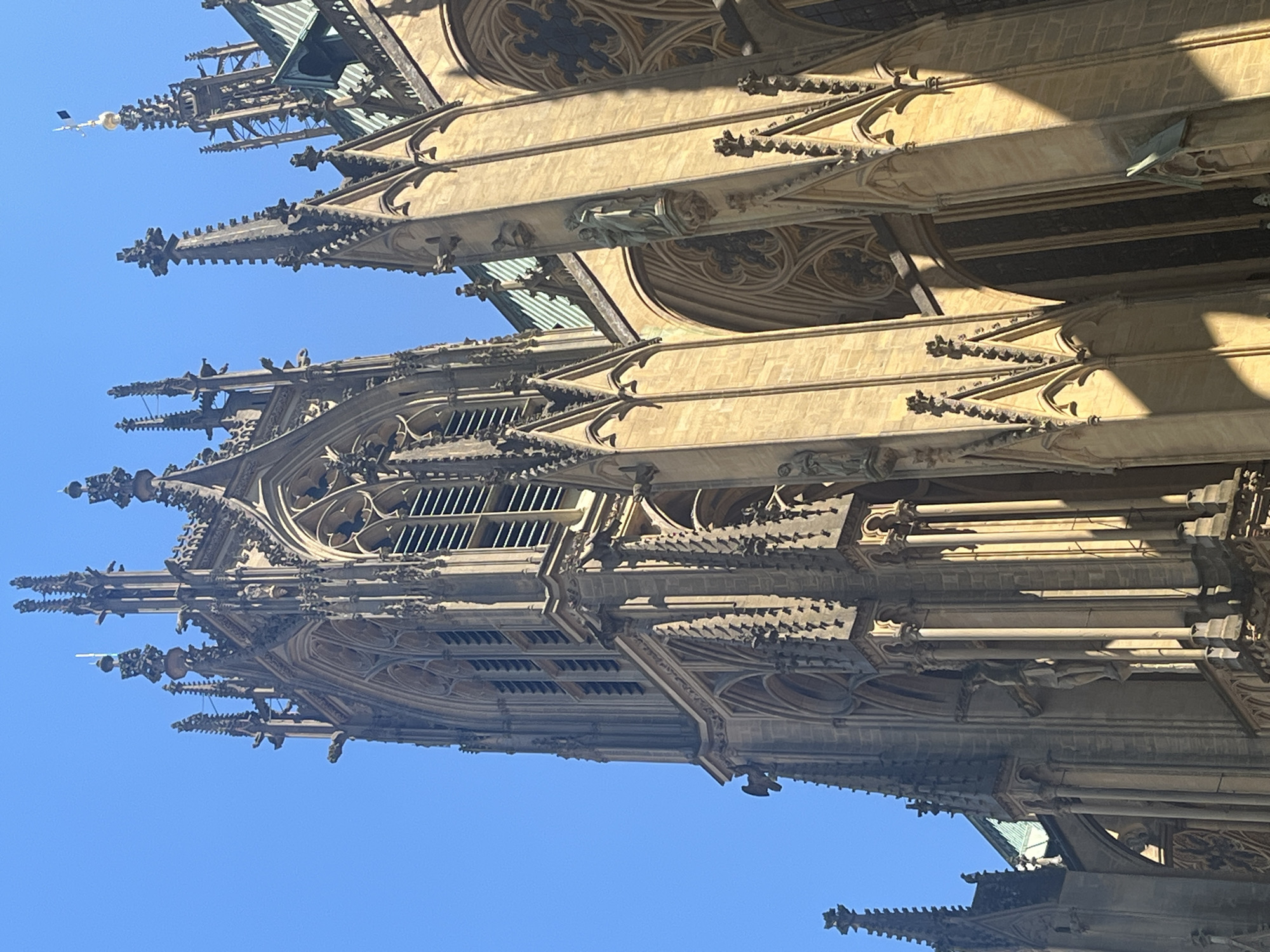 The image captures a section of Metz Cathedral in Metz, France, focusing on its exterior architectural details. The structure exhibits a Gothic style with pointed arches, ribbed vaults, and flying buttresses adorned with decorative elements, including pinnacles and gargoyles. The stone facade shows a range of light to medium beige tones due to natural weathering and sunlight. The background is a clear blue sky. There are no people or immediate surroundings visible. The angle of the shot is upward, accentuating the height and complexity of the cathedral's design.