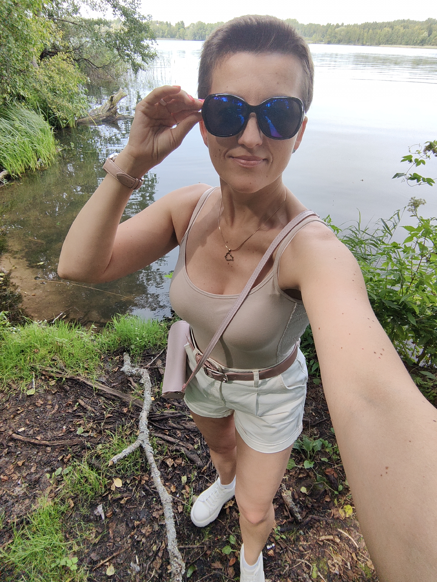 A woman with short brown hair and sunglasses stands on the shore of a lake in Мядзельскі сельскі Савет, Belarus. She is wearing a beige tank top, white shorts, a brown belt, and white sneakers. She is holding her sunglasses with her right hand. A pink purse is slung over her shoulder. The background shows trees and a body of water. The ground is covered with dirt, grass, and fallen branches.