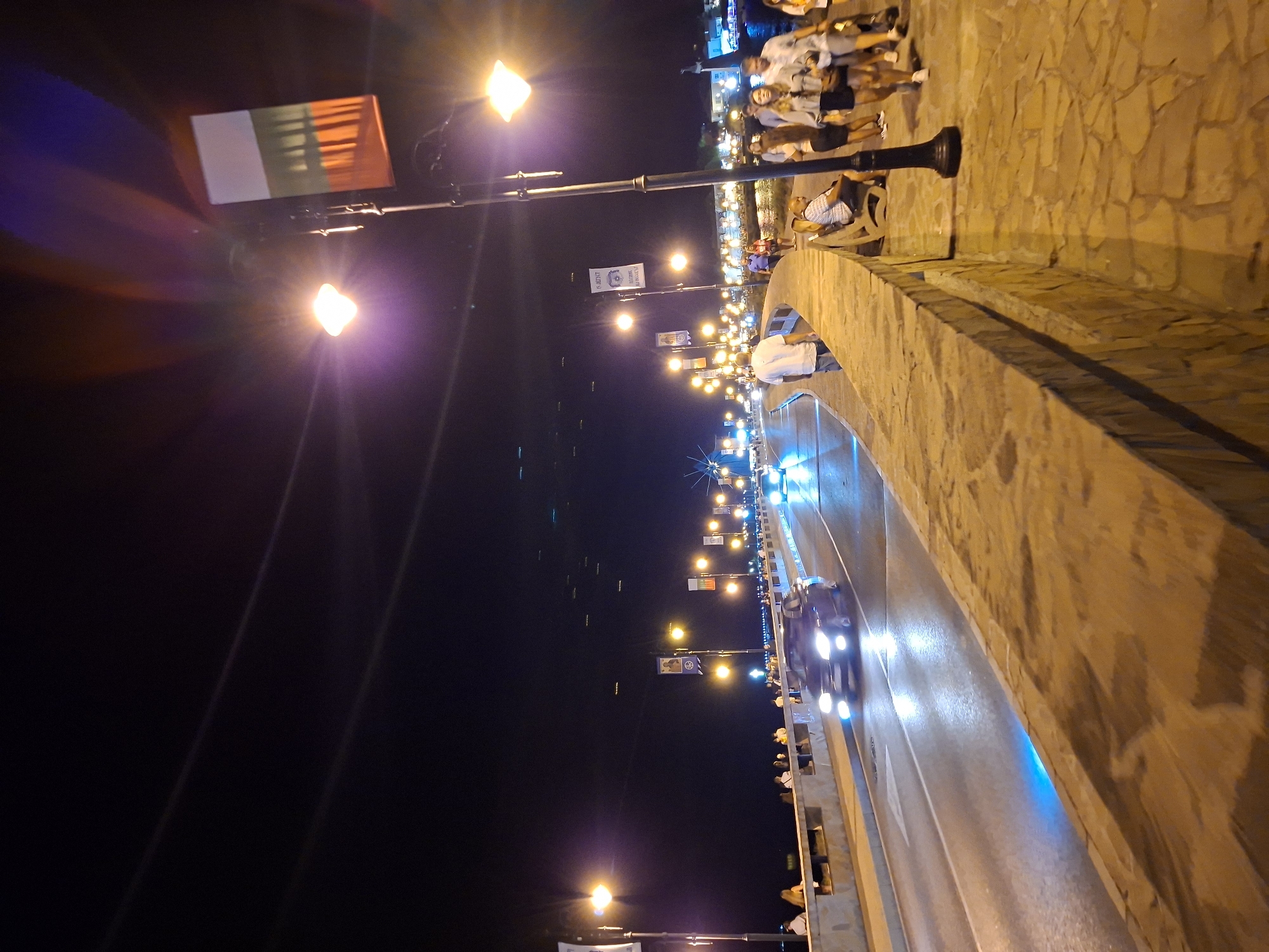 This is a nighttime photograph of a pedestrian walkway and road, likely a bridge or promenade, in an urban setting. The scene is illuminated by streetlights and the headlights of passing vehicles.
On the left side of the image, a road with two lanes of traffic is visible. Several cars are driving on the road, with their headlights on. One car in the foreground is dark-colored and appears to be moving away from the viewer. Another car further down the road is lighter in color and its headlights are also illuminated. The road surface is wet and reflects the lights. Blue lighting is emanating from the side of the road in the foreground, creating an illuminated strip on the asphalt.
The pedestrian walkway is on the right side of the road, separated by a stone wall. The walkway is paved with irregular stones and is lined with several streetlights. A number of people are walking or standing on the walkway. In the foreground, a group of people are walking towards the right side of the image. One man in a white shirt is walking away from the viewer on the walkway. A man is sitting on a bench. Further down the walkway, more people are visible, some walking and some standing.
Above the walkway, several banners are hanging from lampposts. One banner in the upper right corner shows the colors of a flag: white, green, and red, with vertical stripes. Other banners, featuring what appear to be portraits and text, are also visible.
The background is dark, indicating nighttime. The sky is black, with some visible light streaks likely caused by the camera's exposure to bright lights. In the distance, blurred lights suggest buildings or structures. The overall atmosphere is lively, suggesting a popular area for evening strolls or gatherings.