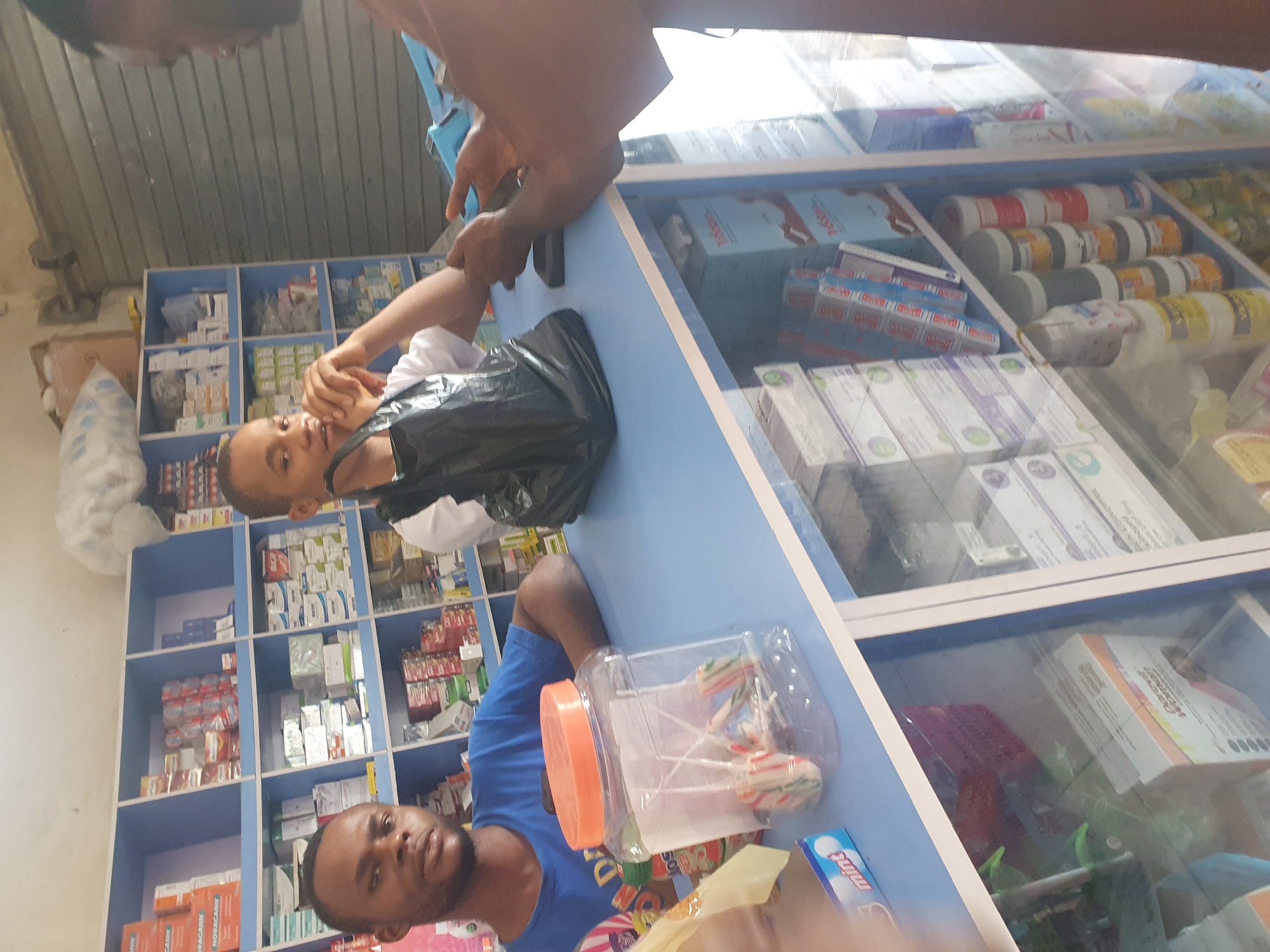 Two men are in a pharmacy in Gwagwalada, Nigeria. Shelves stocked with pharmaceuticals are visible behind them. One man wears a blue shirt. Another wears a white shirt and carries a black bag. A plastic jar filled with lollipops sits on the counter in front of them. The pharmacy display case is filled with medications.