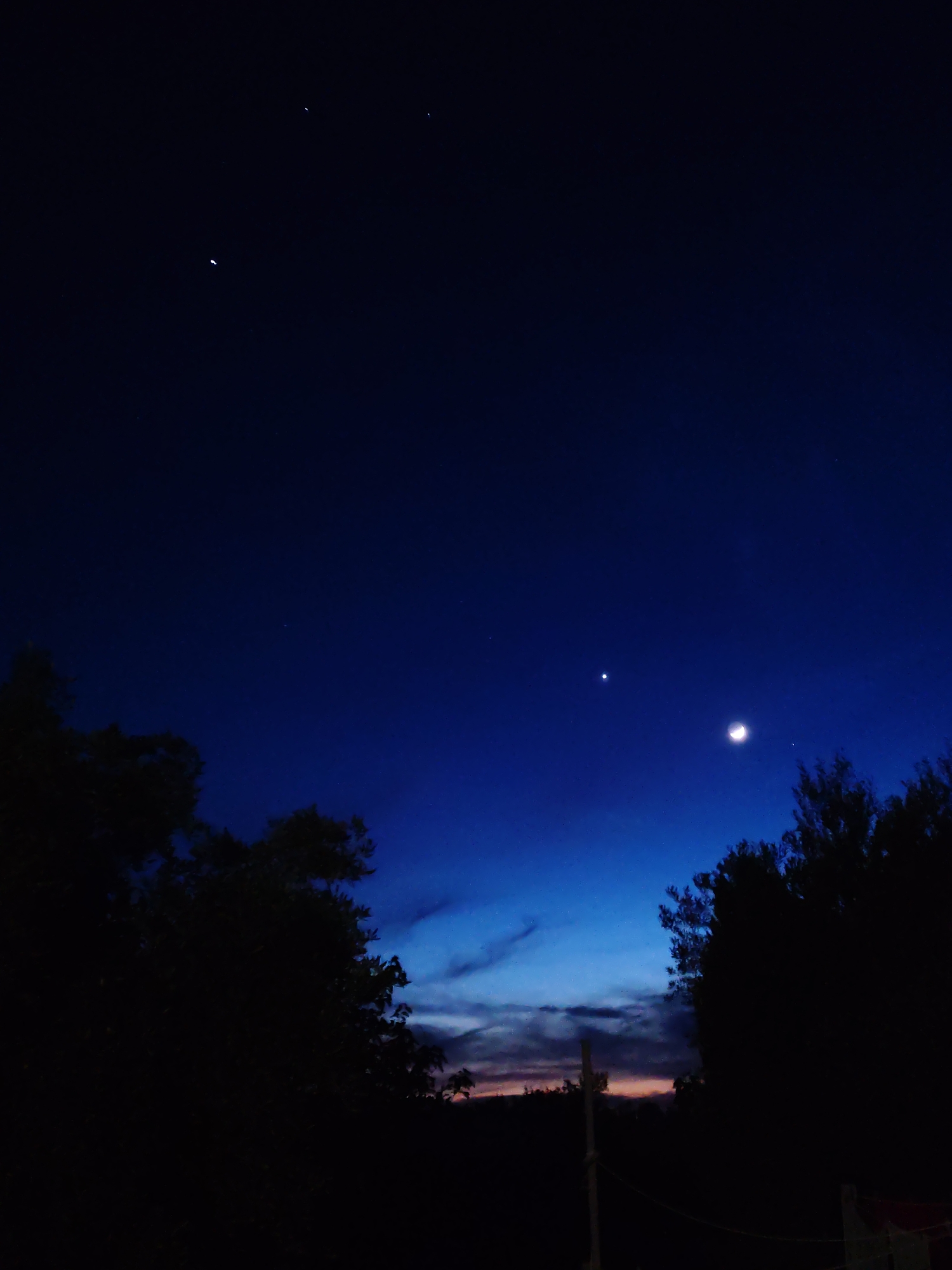 The image depicts a night scene with a dark blue sky. A crescent moon is visible, accompanied by a bright celestial object, possibly a planet, slightly to the left. Several faint stars are scattered across the upper portion of the sky. Silhouetted trees occupy the lower part of the frame, obscuring the landscape, with hints of a horizon line in the center between the trees. The location is unknown.