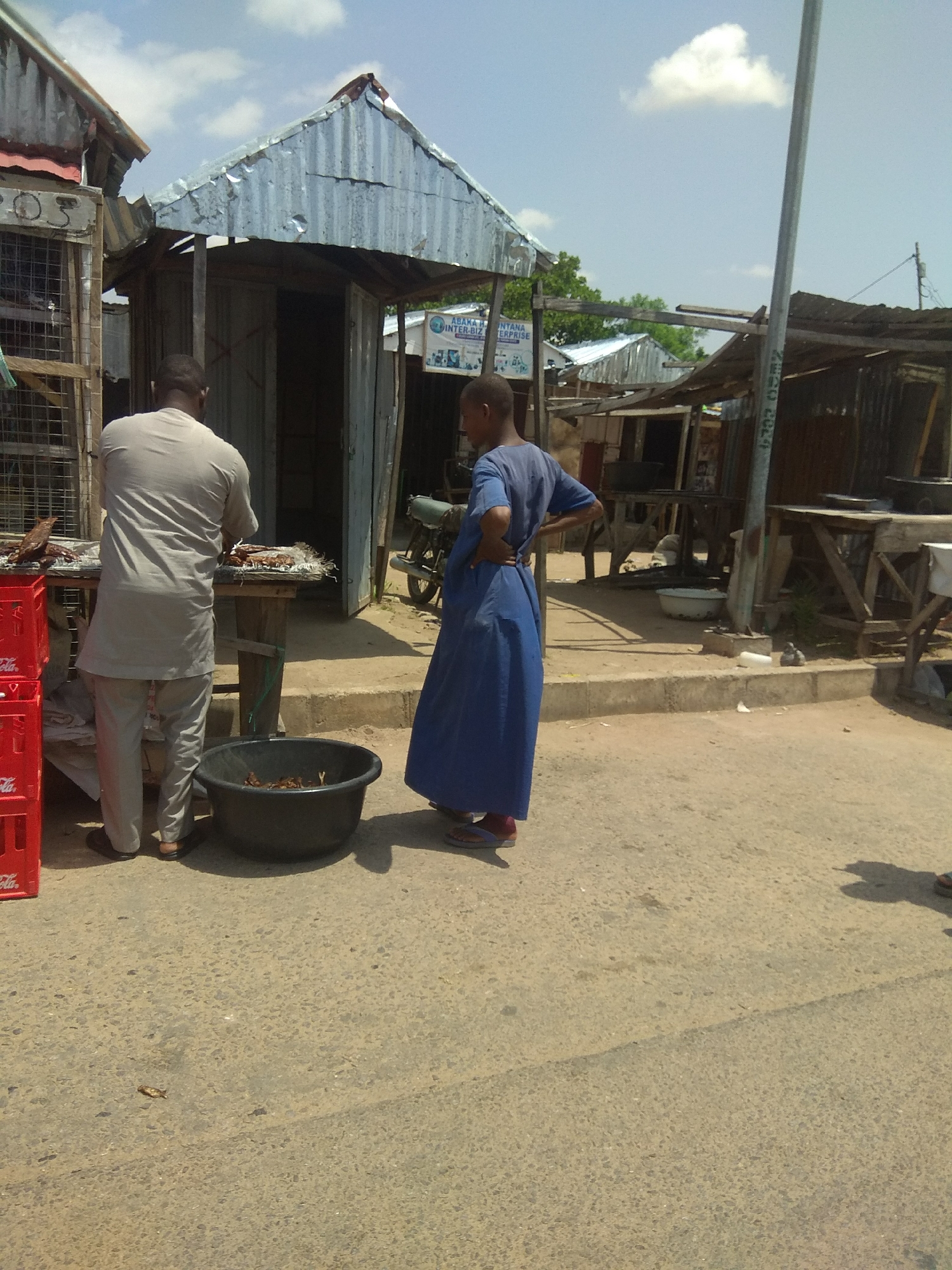 In Jalingo, Nigeria, on a sunny day with scattered clouds, two individuals stand near makeshift market stalls constructed from corrugated metal and wood. To the left, a man in a light gray tunic and trousers faces a wooden table laden with dried fish. He appears to be arranging or preparing the fish for sale. To his right, a younger person in a long blue garment stands with hands on their hips, observing. A large black basin filled with dried fish sits on the ground in front of the man. Red crates, possibly for beverages, are stacked to the left of the man. In the background, other market structures and a motorcycle are visible. The ground is paved with asphalt. The overall scene depicts a typical street market environment in Jalingo.
