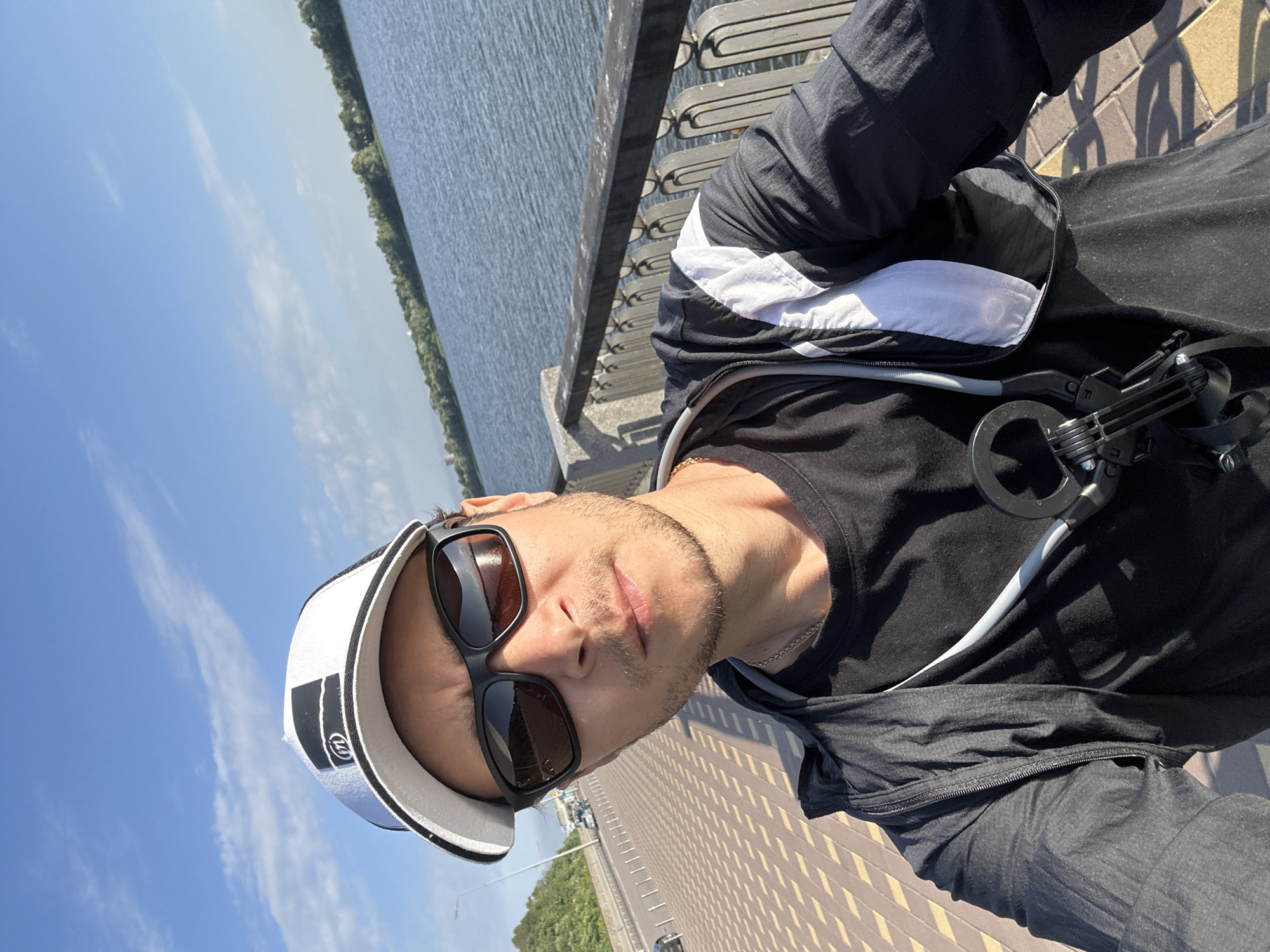 A man is taking a selfie outdoors on a sunny day. He is wearing a white baseball cap with black trim and a logo, black sunglasses, a black t-shirt, and a black and white jacket. He has a slight stubble. The man is standing on a paved walkway with a patterned design. To his right, there is a decorative metal fence and a large body of water, likely the Dnipro River, with trees and buildings visible on the opposite bank. In the background to his left, there is a road with cars and a lamppost. The sky is blue with some scattered white clouds. The location appears to be a promenade or embankment in Kyiv, Ukraine.