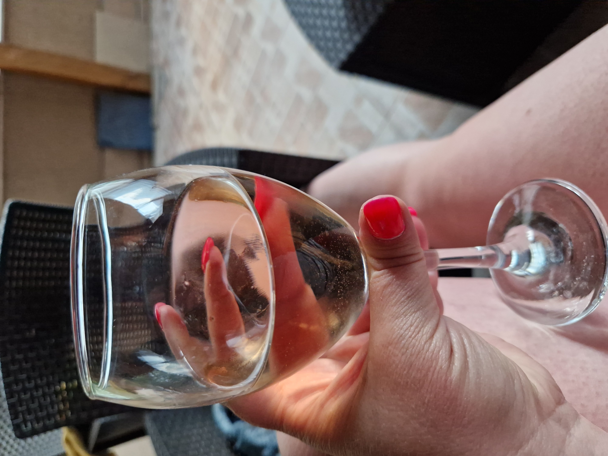 In this close-up shot, a hand with bright red nail polish holds a wine glass filled with a pale, bubbly liquid. The glass is transparent, and the contents appear to be a white wine or sparkling beverage. The hand is positioned to obscure part of the background, which seems to be an outdoor setting with rattan furniture and a tiled floor, suggesting a patio or terrace. The lighting indicates it is daytime. The image does not contain any discernible elements that would place it in a specific city or location.